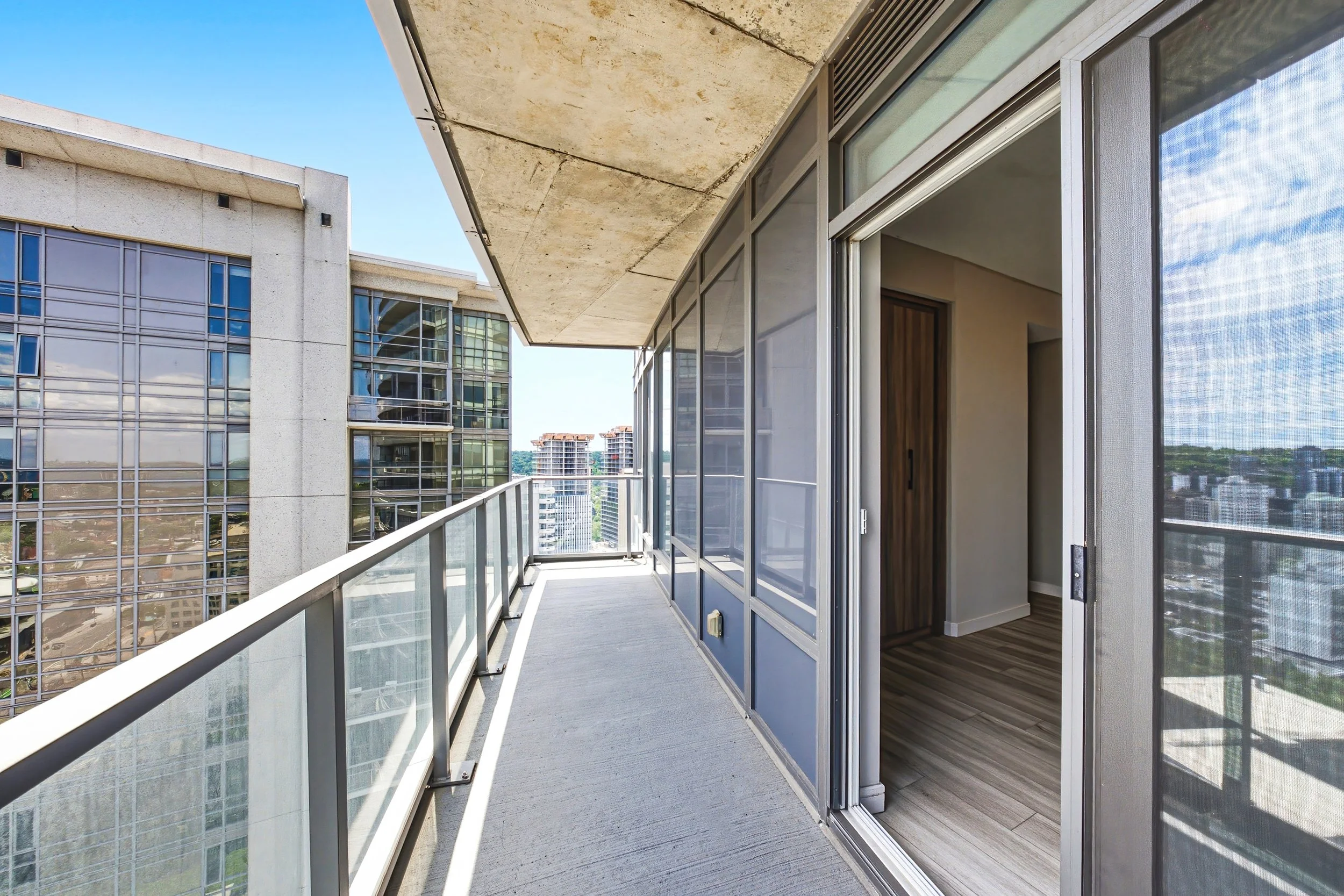 Balcony with glass railing, open sliding glass doors revealing an interior room, and view of other high-rise buildings under clear blue sky.