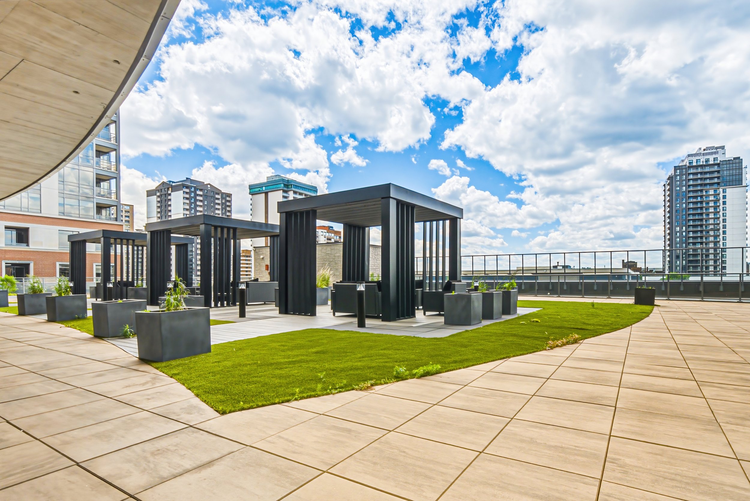 Urban rooftop terrace with black pergolas, planters, and green grass, surrounded by high-rise buildings under a partly cloudy sky.