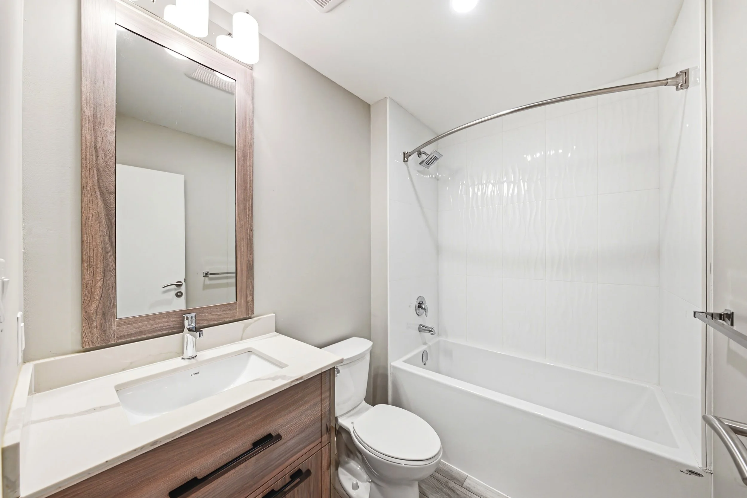 A bathroom with a modern design, including a wooden framed mirror above a white sink, a toilet, and a bathtub with a curved shower rod. The walls are painted light and there is a white tile wall in the bathtub area. The floor appears to be wood or wo