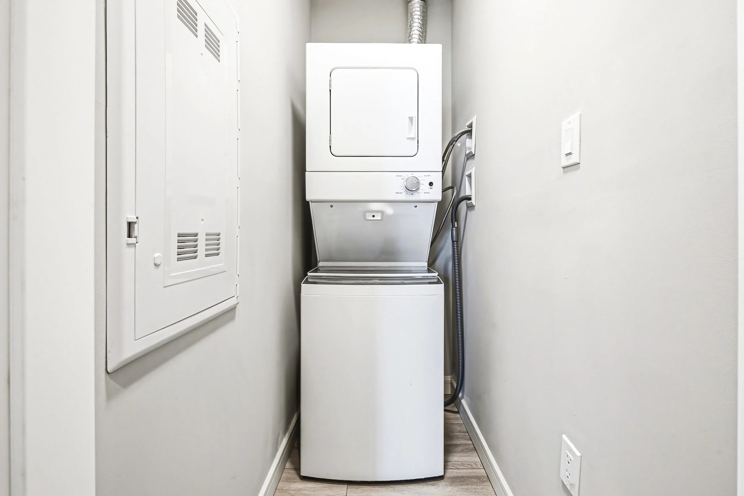 A laundry room with a stacked clothes dryer and washing machine, located between white walls and a white utility cabinet on the left.