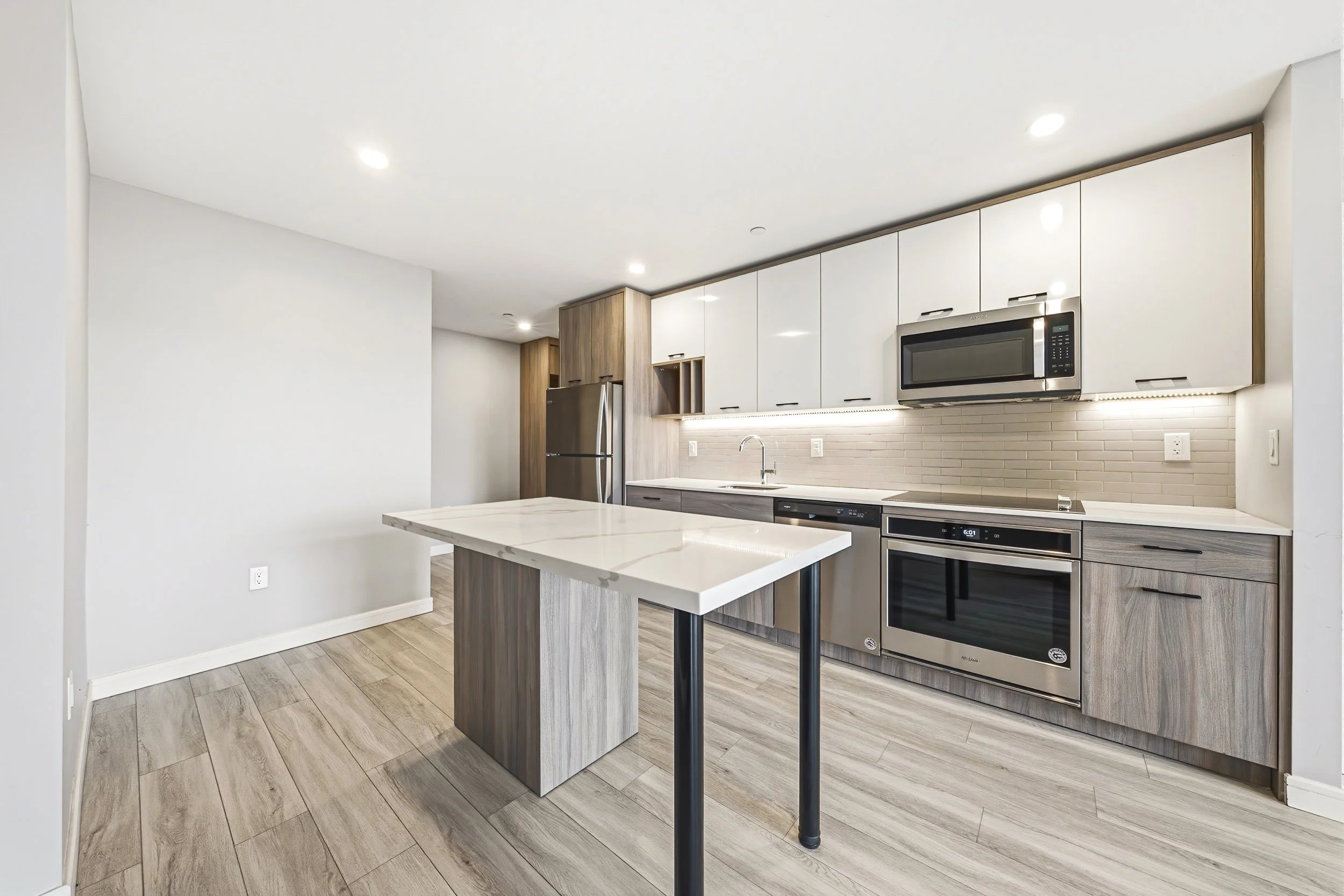 Modern kitchen with white cabinets, a stainless steel microwave, oven, and refrigerator, island counter with a marble top, wood flooring, and recessed ceiling lights.