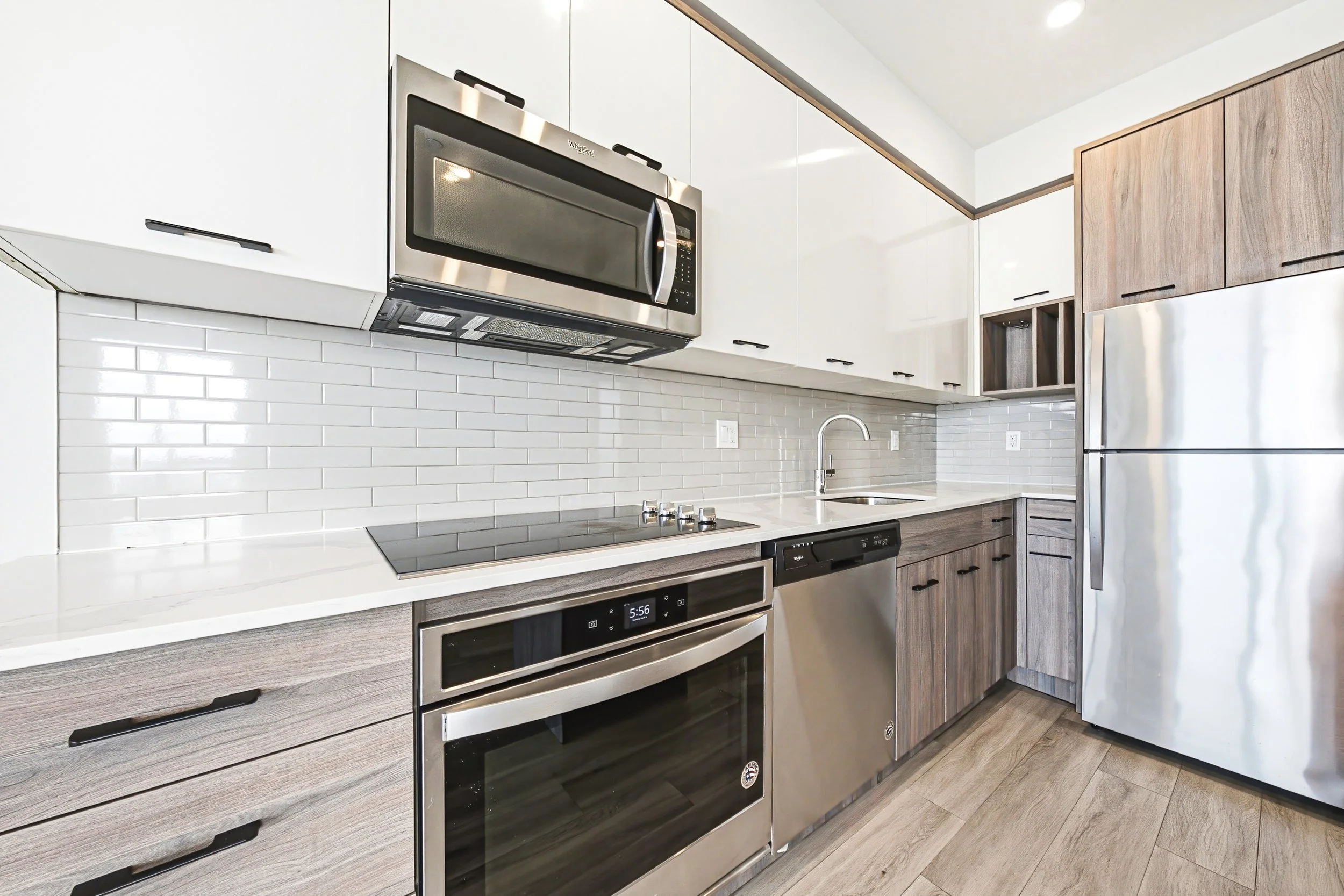 Modern kitchen with white upper cabinets, wood lower cabinets, a stainless steel microwave, oven, and refrigerator, white subway tile backsplash, and a marble countertop.