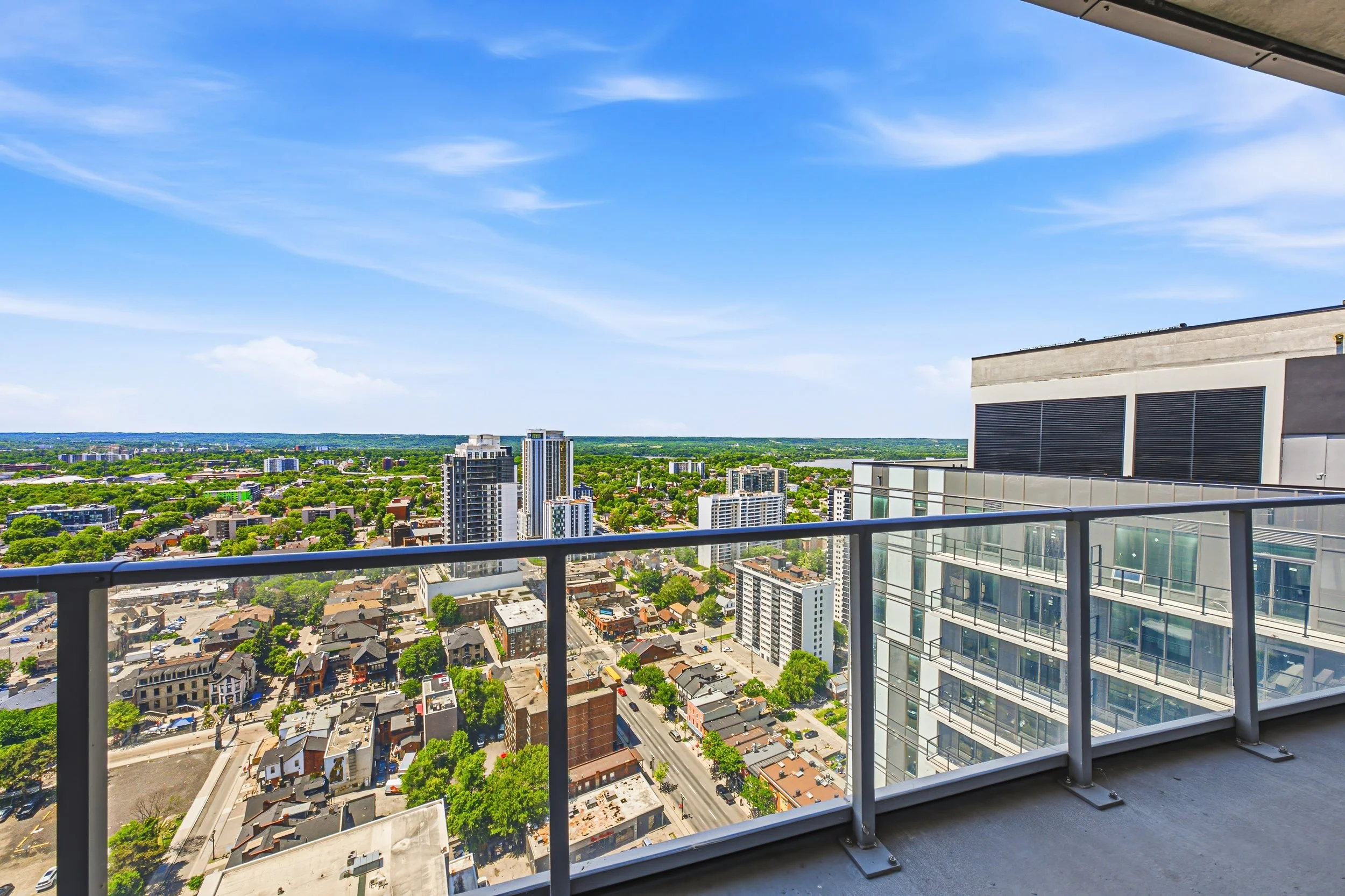 View from a high balcony showing a cityscape with tall buildings, smaller houses, and greenery under a bright blue sky with some clouds.