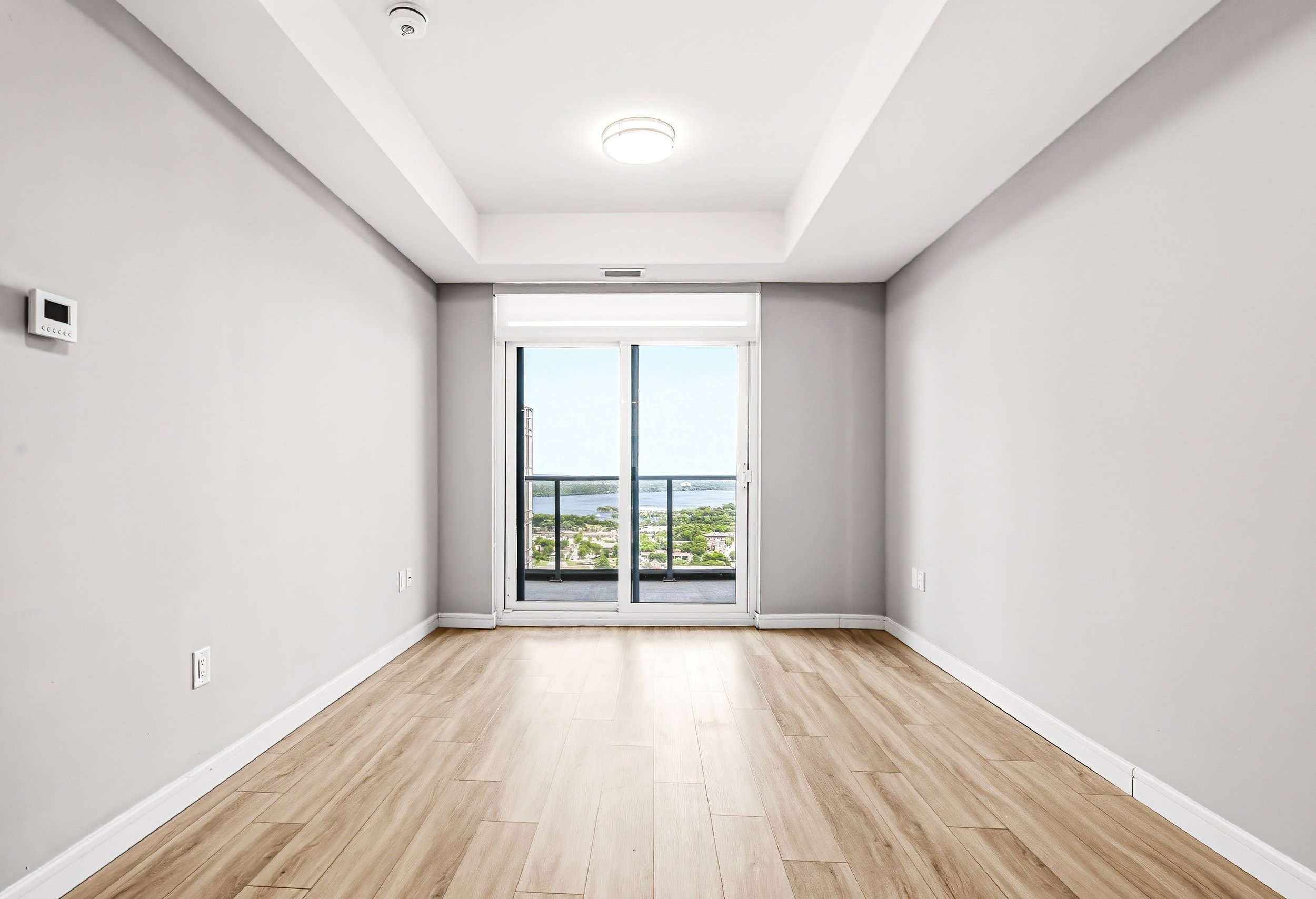 An empty modern room with white walls, light wood flooring, a ceiling light, a thermostat on the wall, and large sliding glass doors leading to a balcony with a view of a river and cityscape.