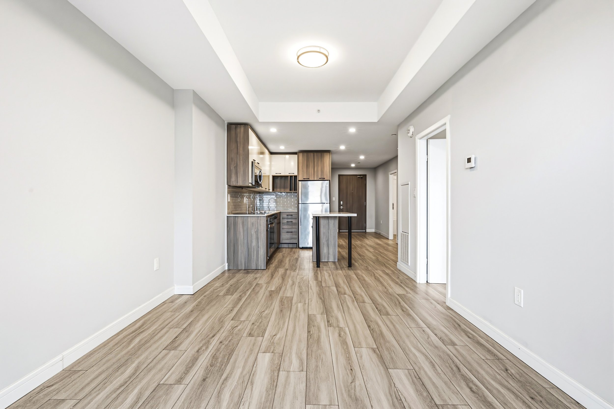 Empty room with light wood flooring, white walls, and an open kitchen with modern cabinetry and stainless steel appliances visible in the background.