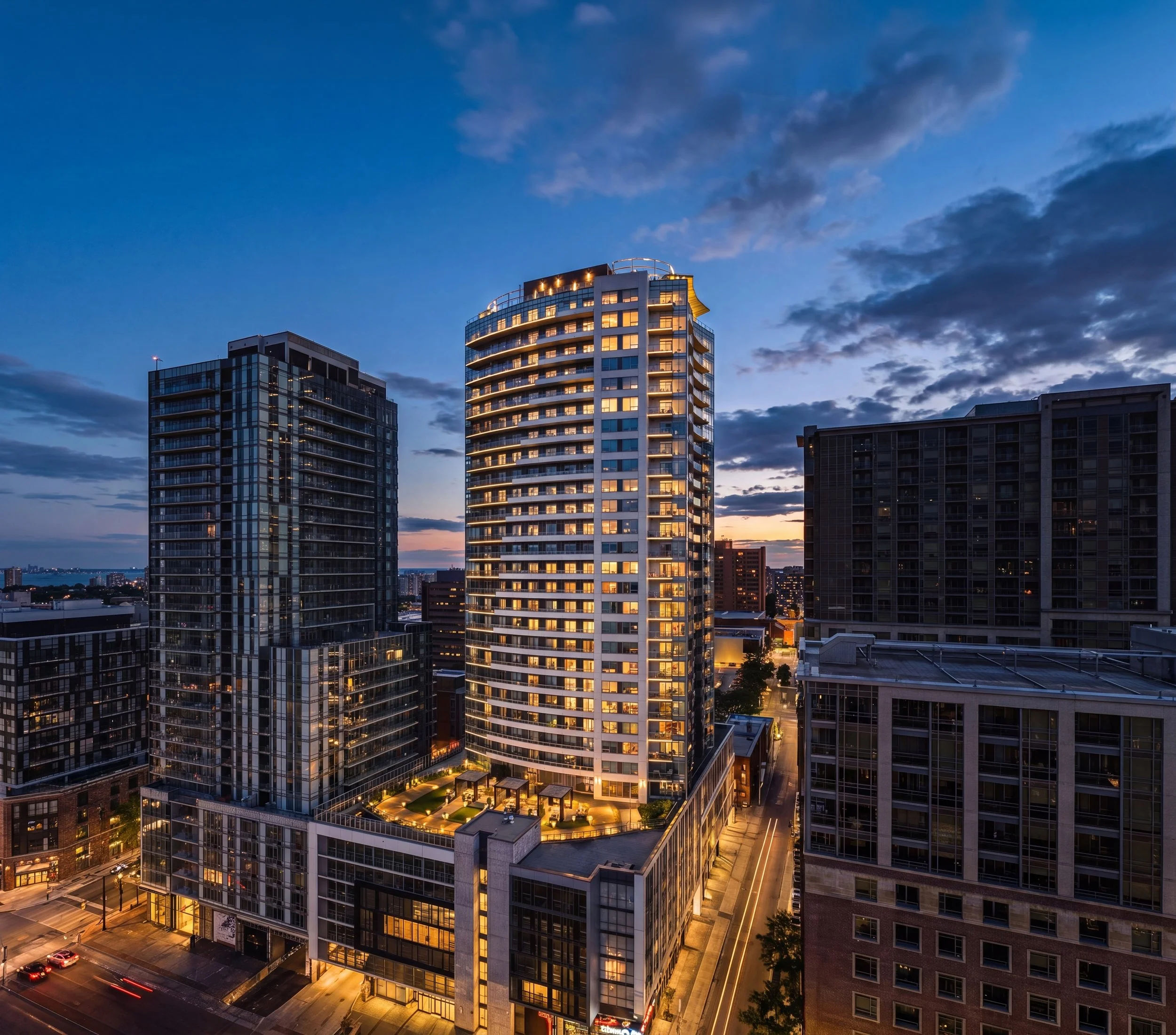 High-rise modern buildings with illuminated windows, street below with cars and light trails, evening sky with clouds and sunset colors.
