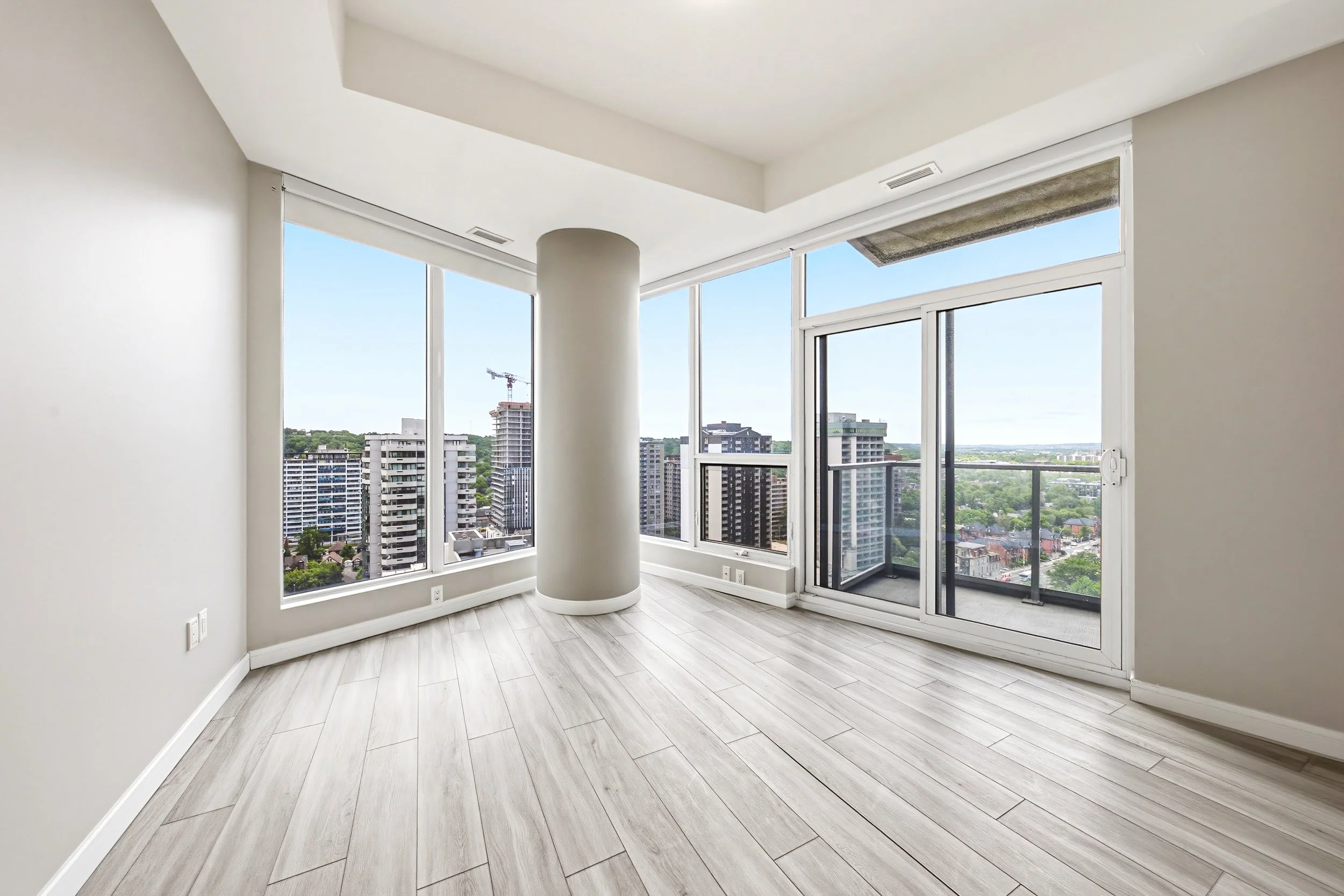 Empty modern apartment room with large floor-to-ceiling windows showing city buildings and a blue sky, white walls, light wooden floor, and a balcony door.
