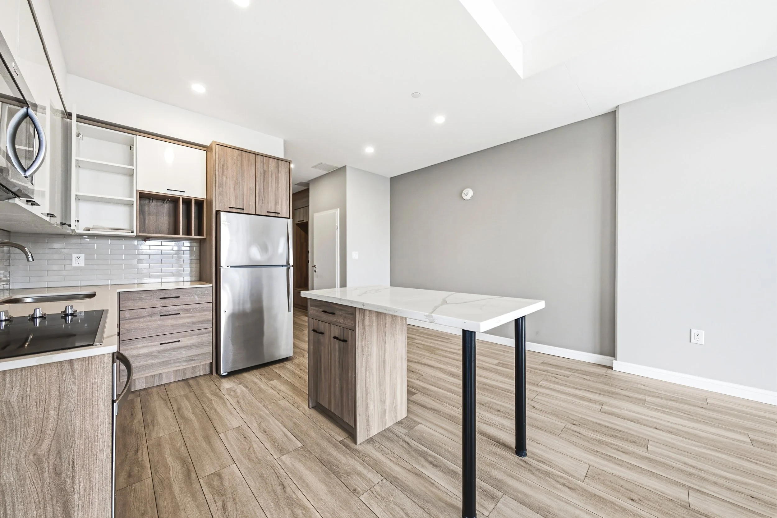 Modern kitchen with light wood cabinets, stainless steel refrigerator, white marble island, and wood flooring, with white walls and ceiling.