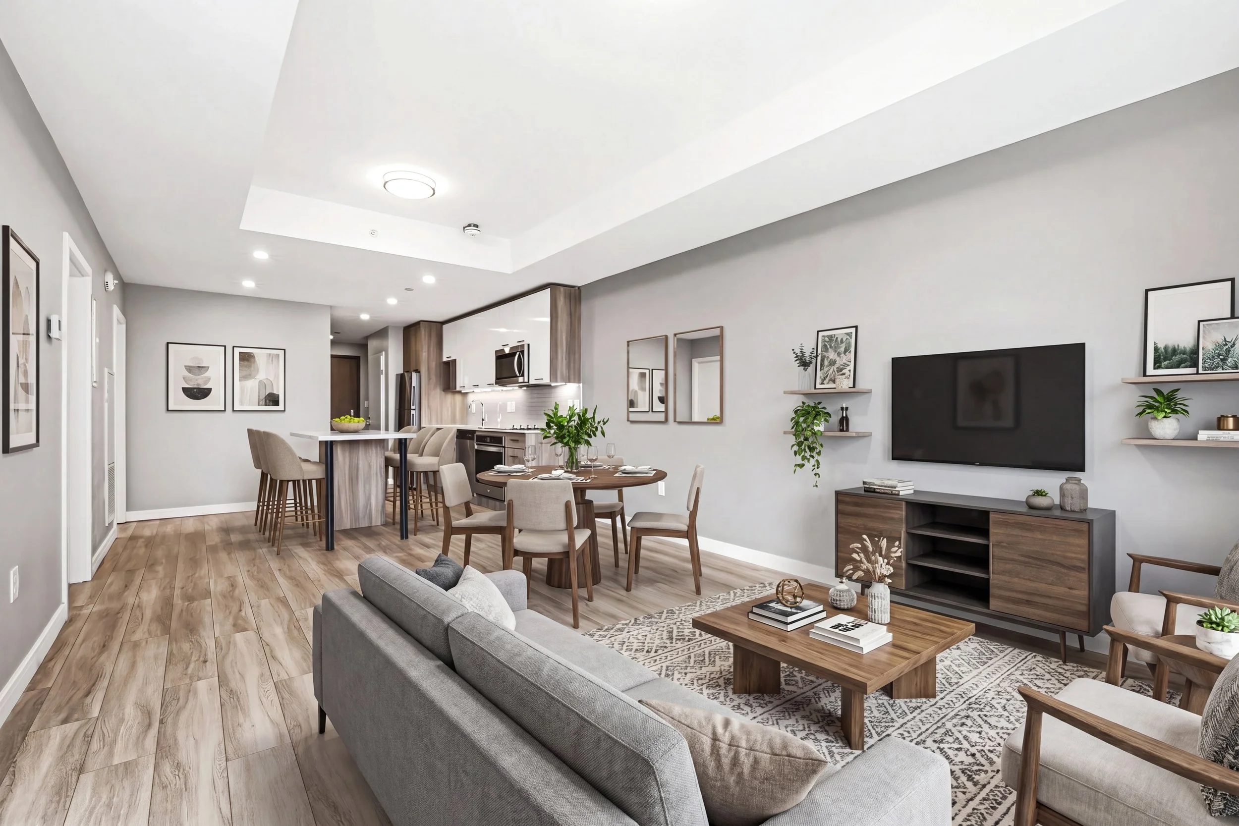 Modern living room with open kitchen, dining area, and decor, featuring wood flooring, gray sofa, dining table with chairs, wall art, plants, and a large flat-screen TV.