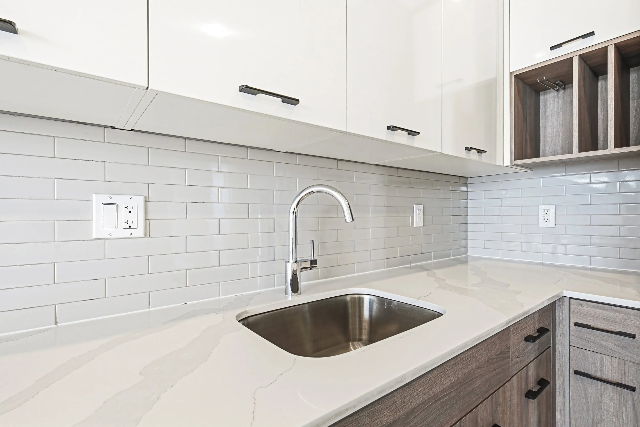 Modern kitchen with white cabinetry, white subway tile backsplash, and a white marble countertop. There is a stainless steel sink with a chrome faucet, electrical outlets on the wall, and open shelving in a wood finish.