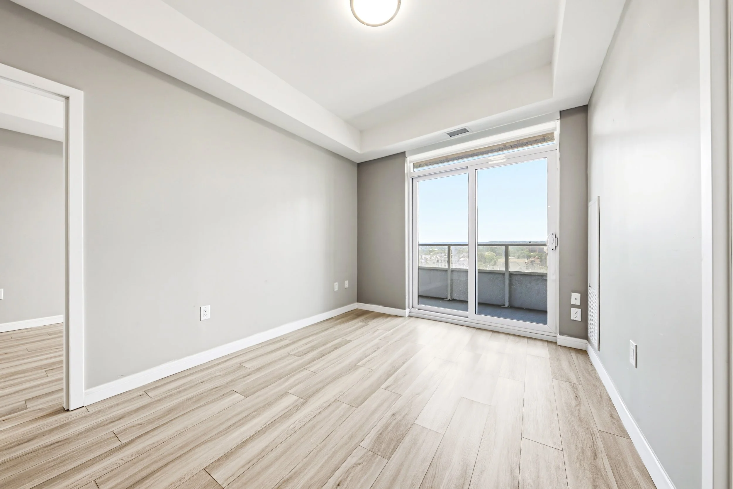 Empty living room with light wood flooring, gray walls, large sliding glass door leading to balcony, ceiling light fixture, and electrical outlets.