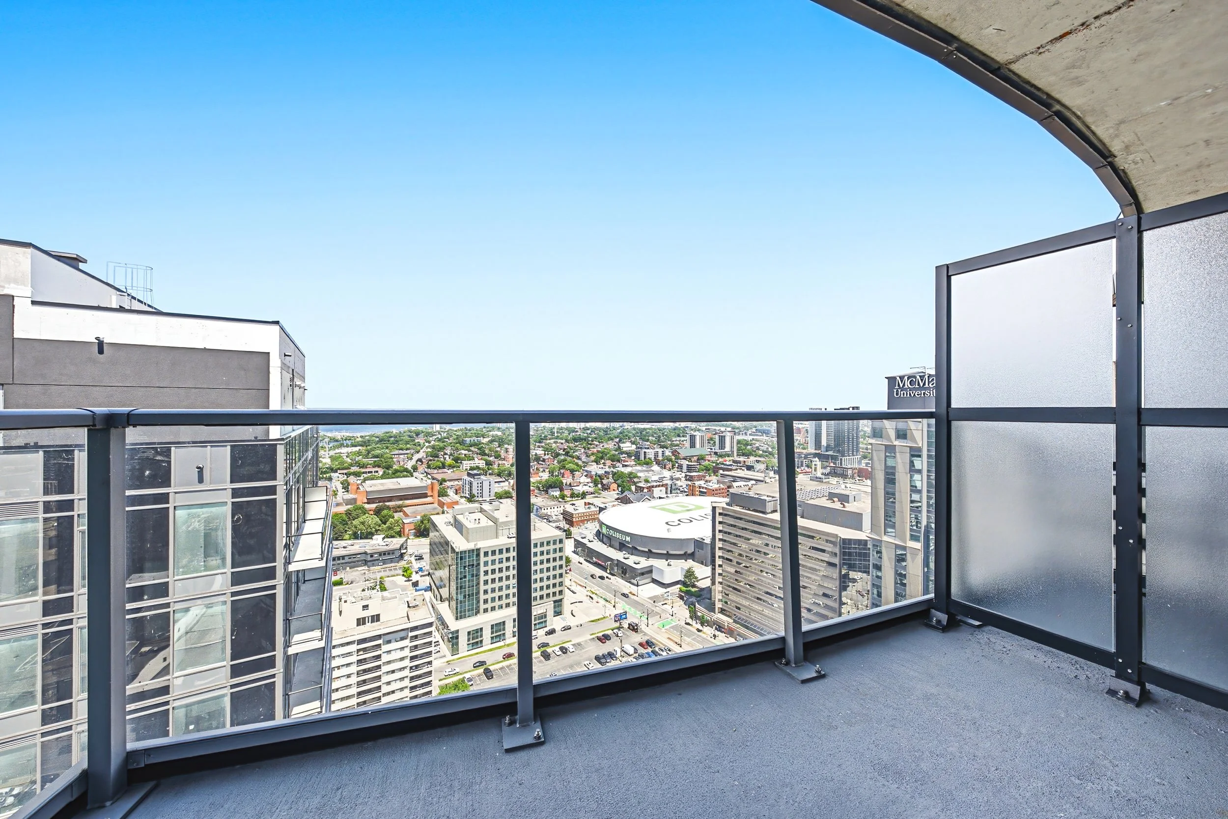 View from a high-rise balcony showing cityscape with buildings, streets, and green areas under a clear blue sky.