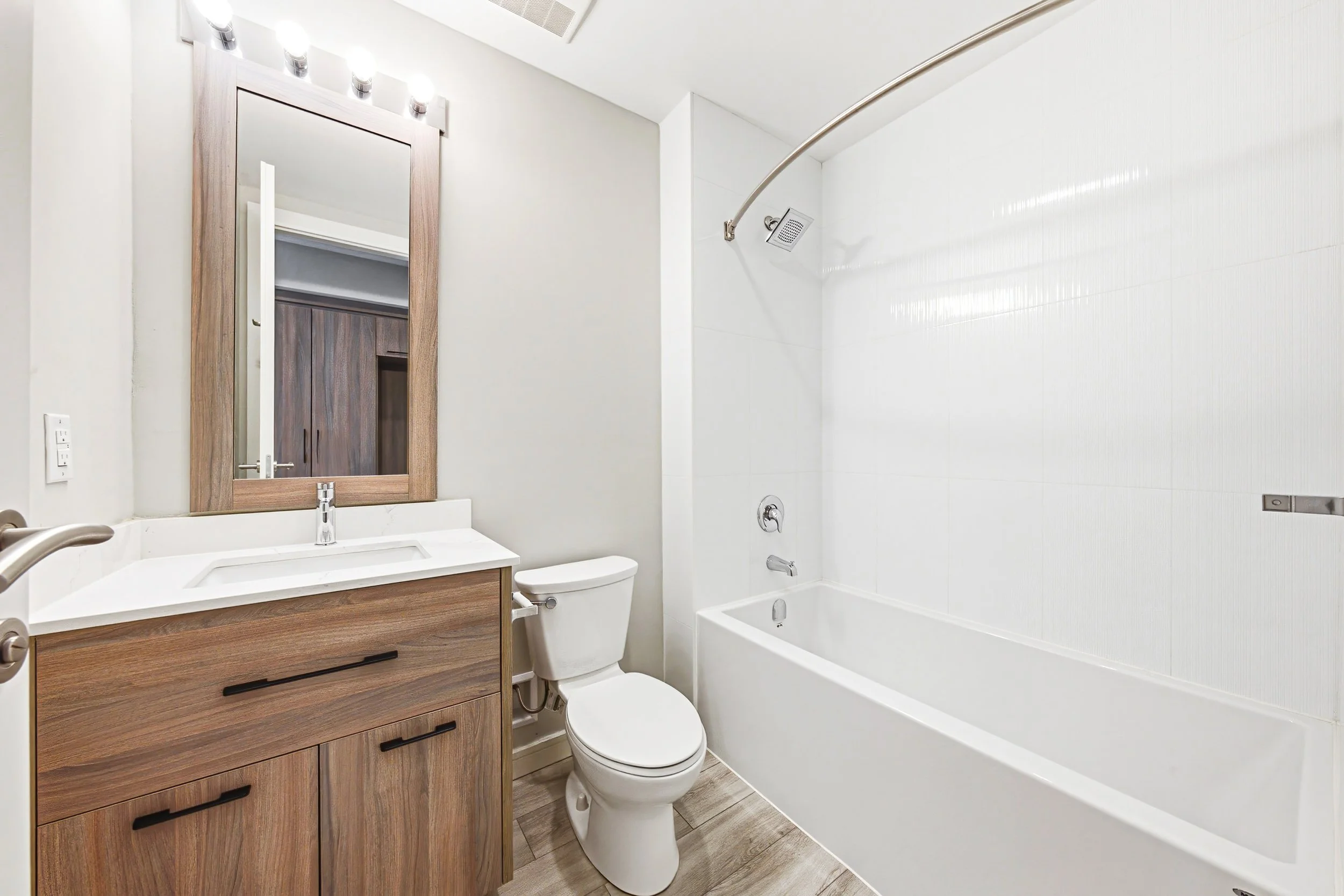 A modern bathroom with a wooden vanity featuring a white sink and black handles, a mirror above the sink, a white toilet, and a bathtub with a curved shower rod and a square shower head, all with white tiled walls and wood-look flooring.