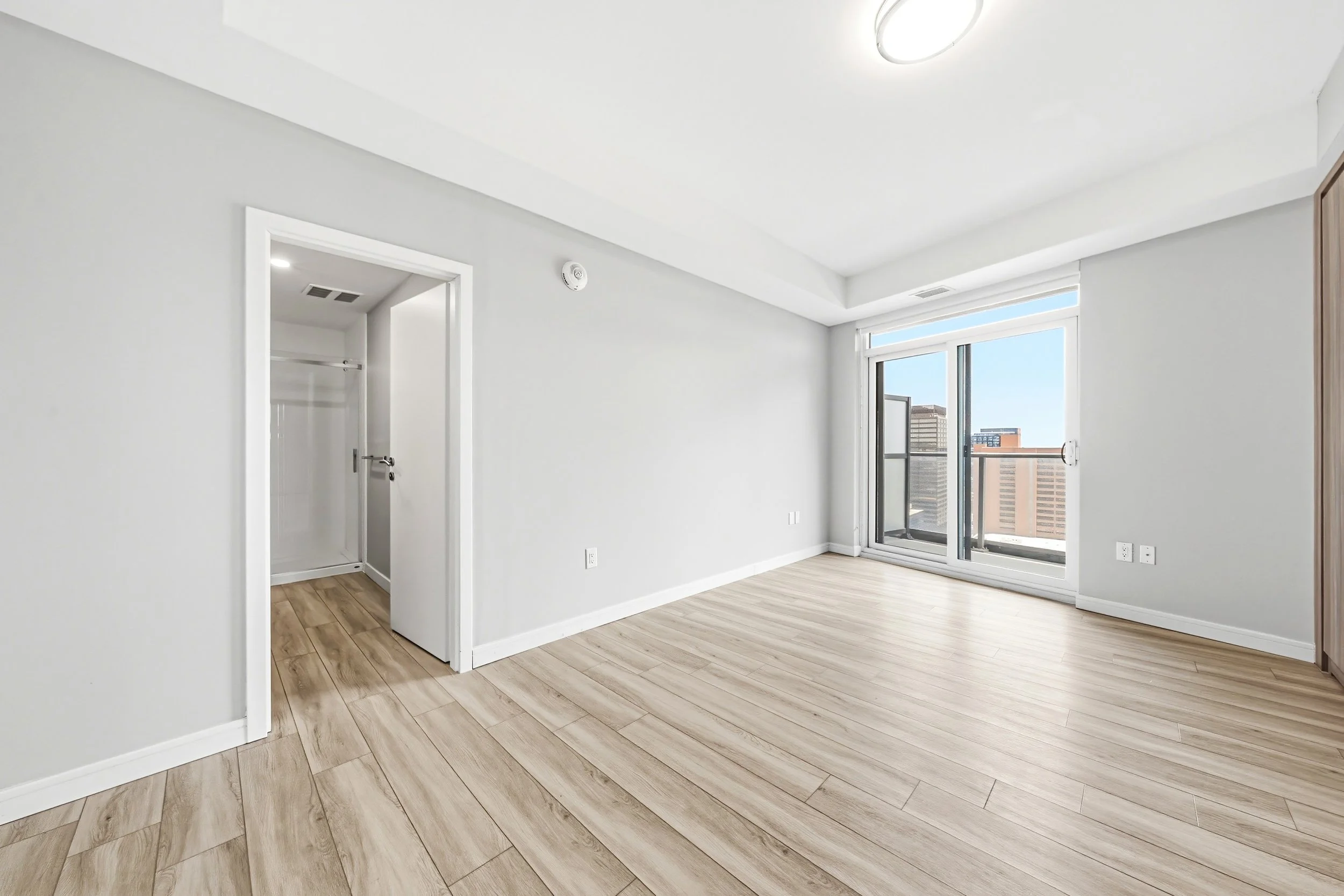 Empty modern apartment room with wood flooring, white walls, and large sliding glass door leading to a balcony with city view.