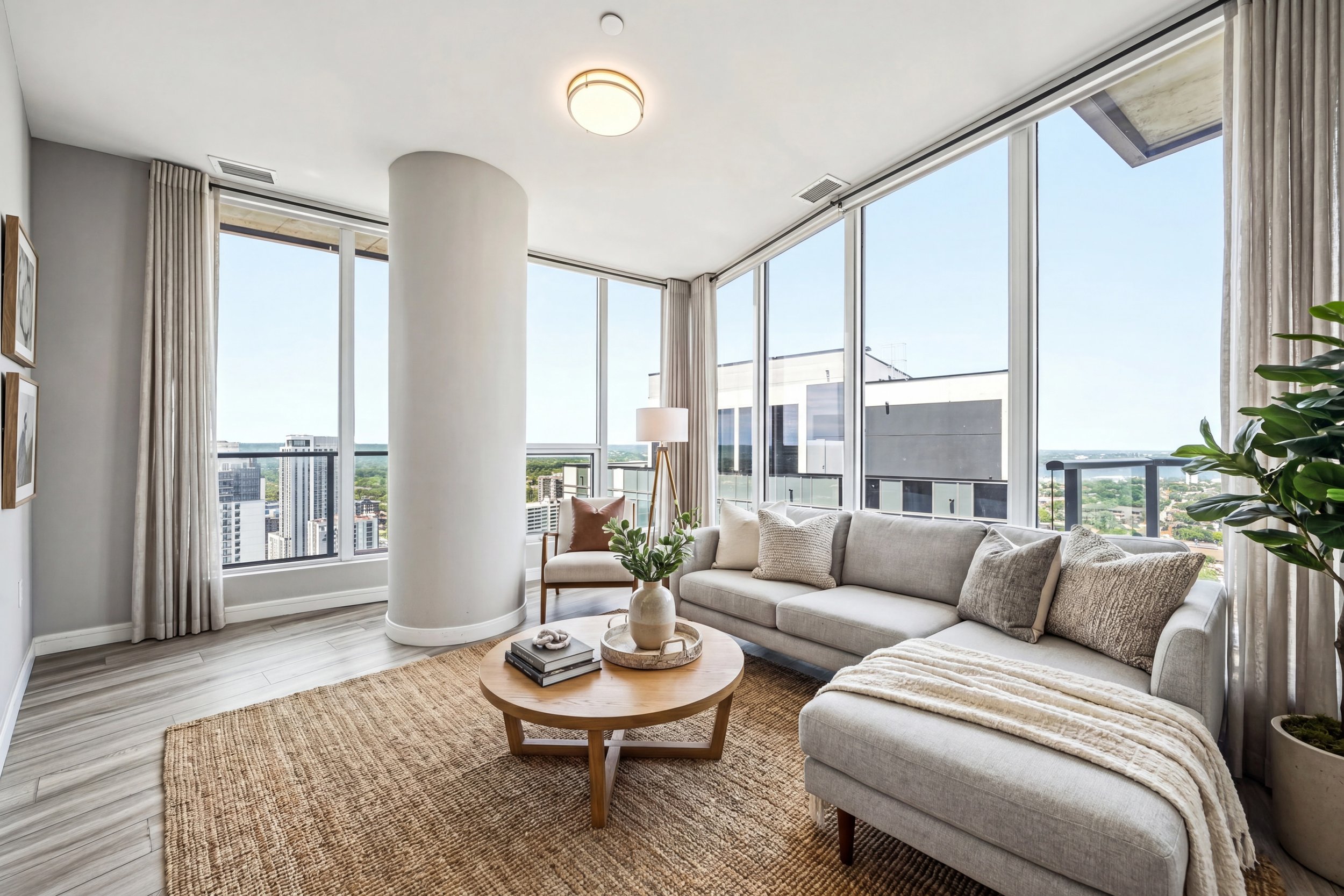 Bright living room with large floor-to-ceiling windows, white sofa with neutral pillows, a round wooden coffee table with books and decor, potted plant, and cityscape view