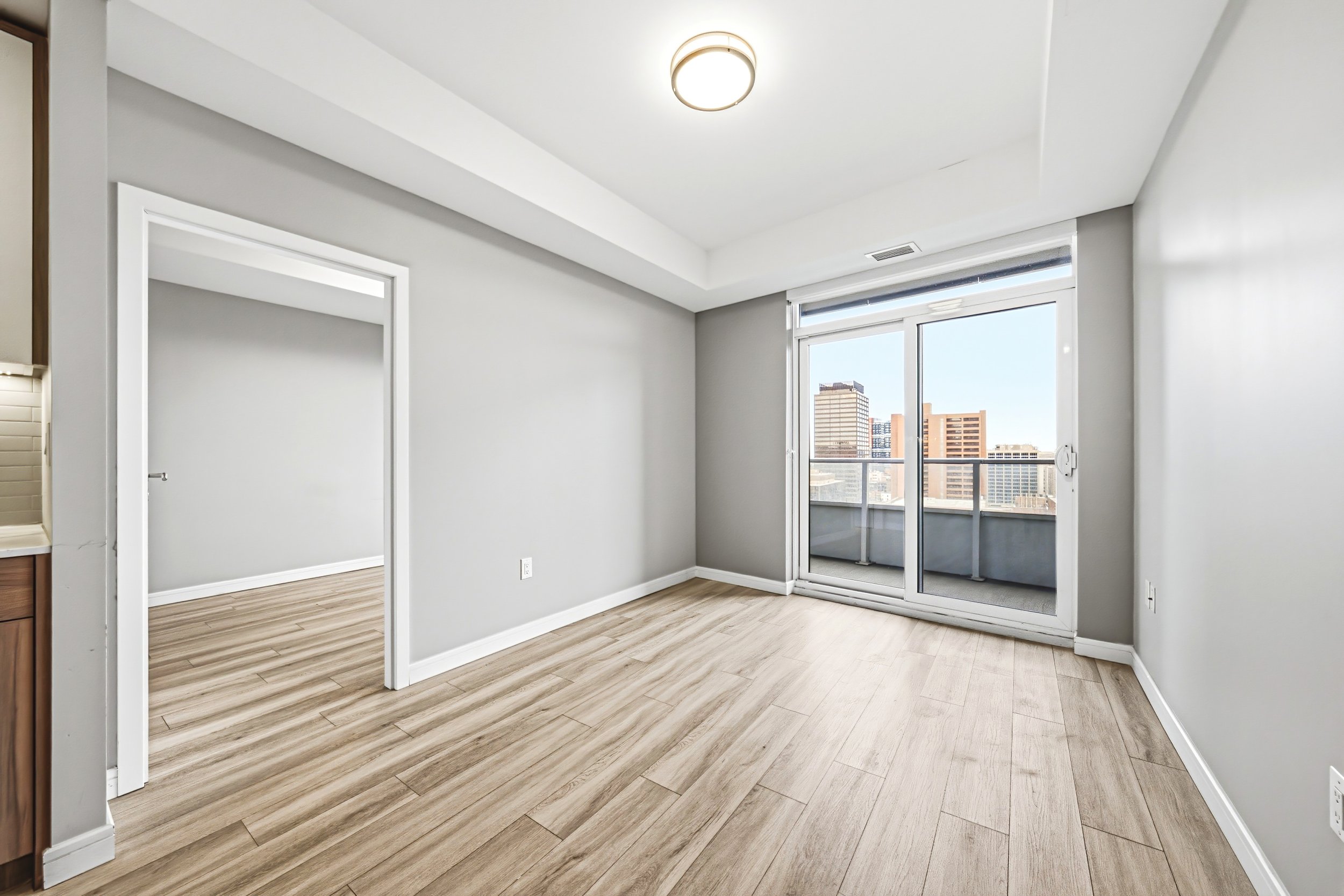 Interior of an unfurnished apartment room with a large window and sliding glass door leading to a balcony, with wood-look flooring and white walls.