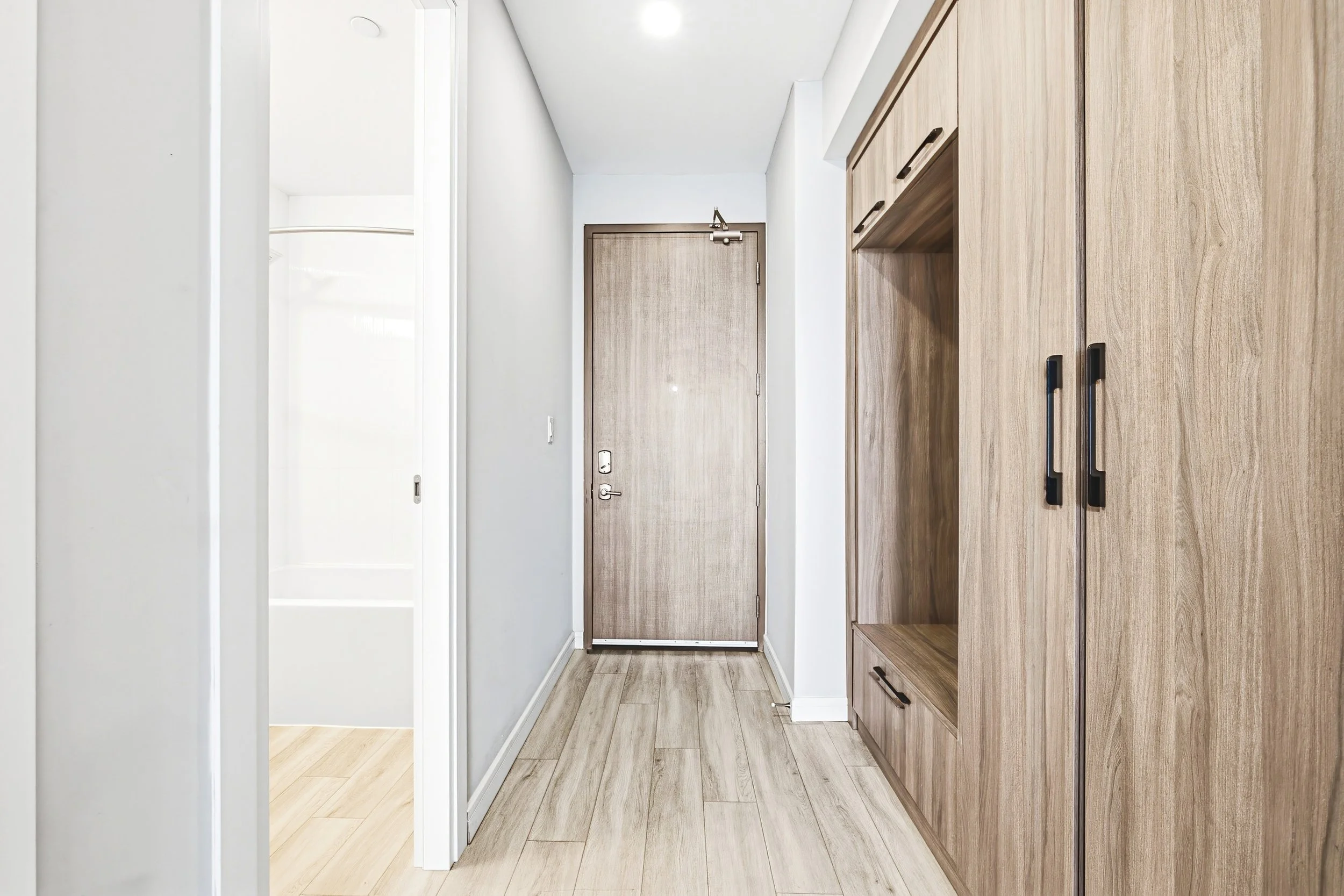 A modern hallway with wooden cabinetry on the right side, a tan front door at the end, white walls, and light-colored wood flooring.