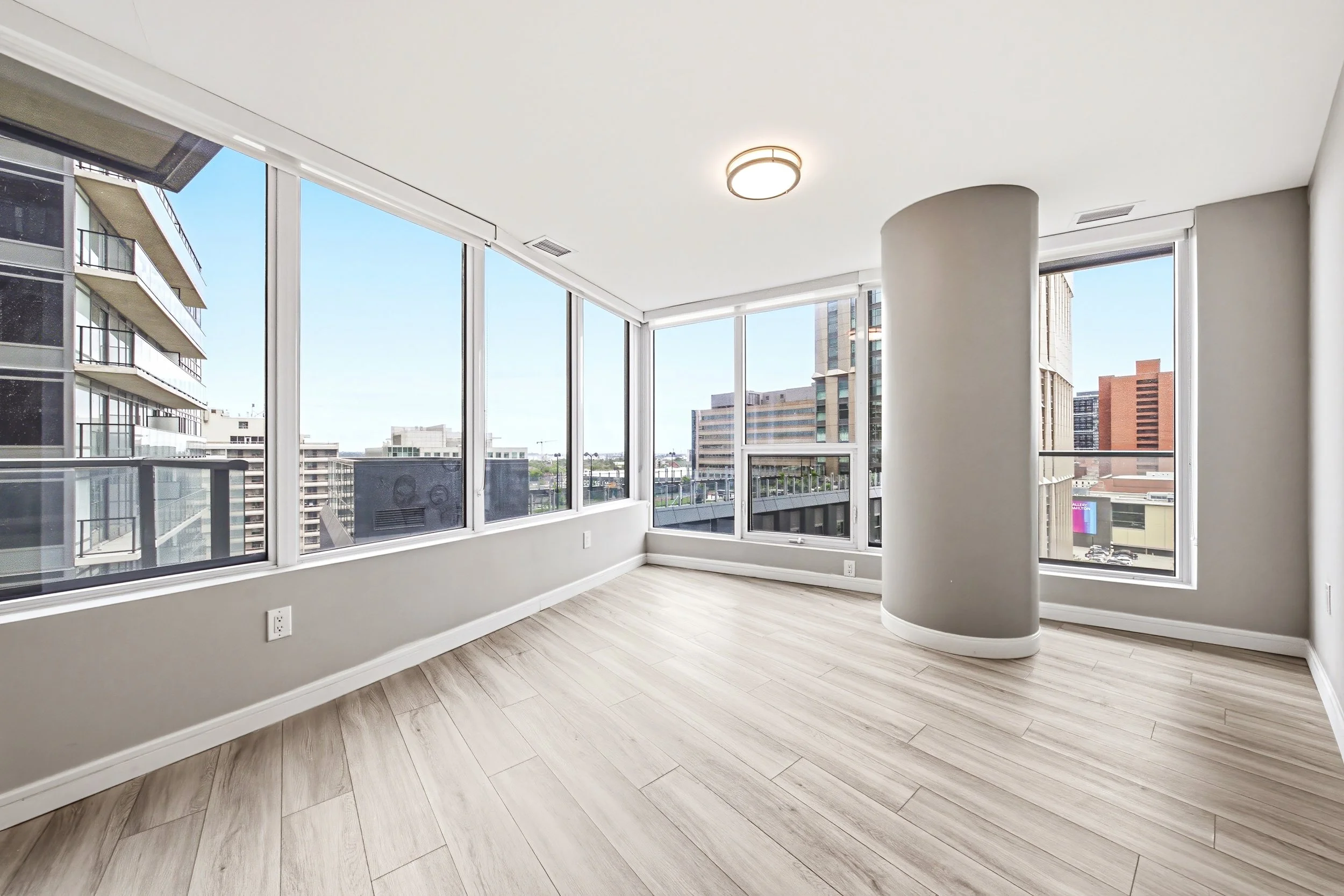 Empty modern apartment room with large floor-to-ceiling windows showing city buildings and blue sky, light wood flooring, white walls, and a circular ceiling light fixture.
