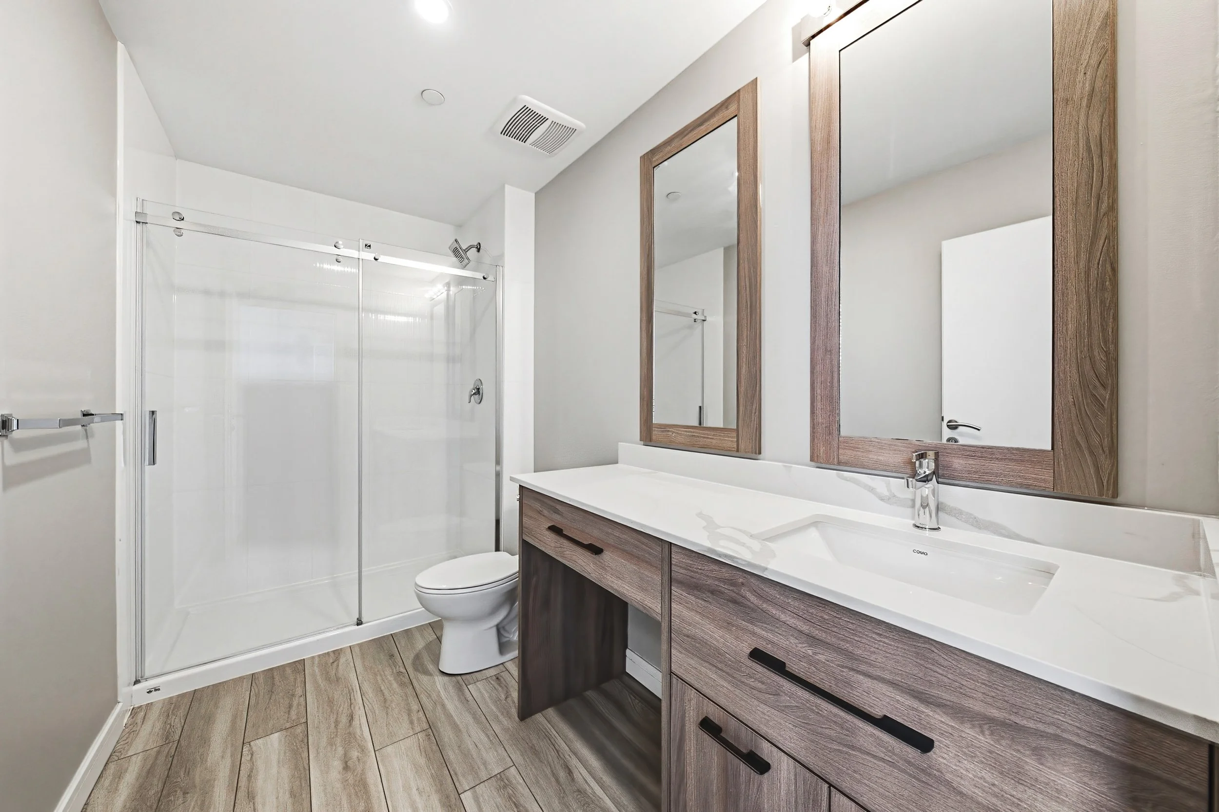 Modern bathroom with wooden vanity, large mirror, sink, toilet, and glass-enclosed shower with sliding doors.