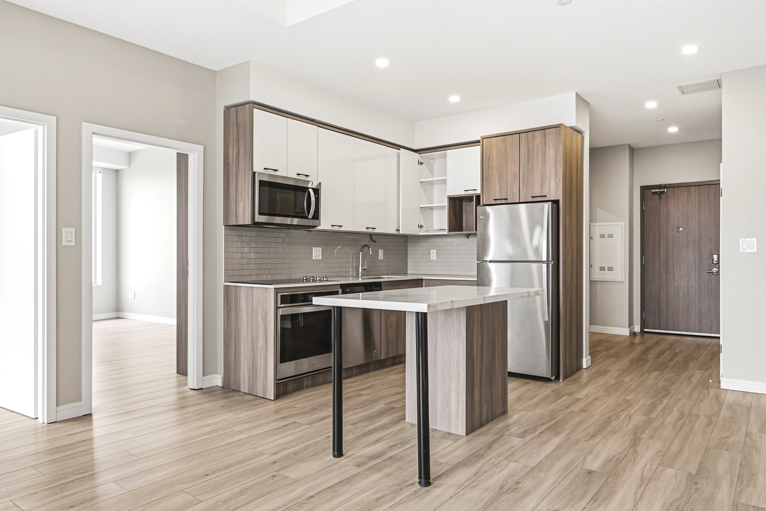Modern kitchen with white and wood cabinets, stainless steel appliances including a microwave and refrigerator, a kitchen island with a marble top, and hardwood flooring.