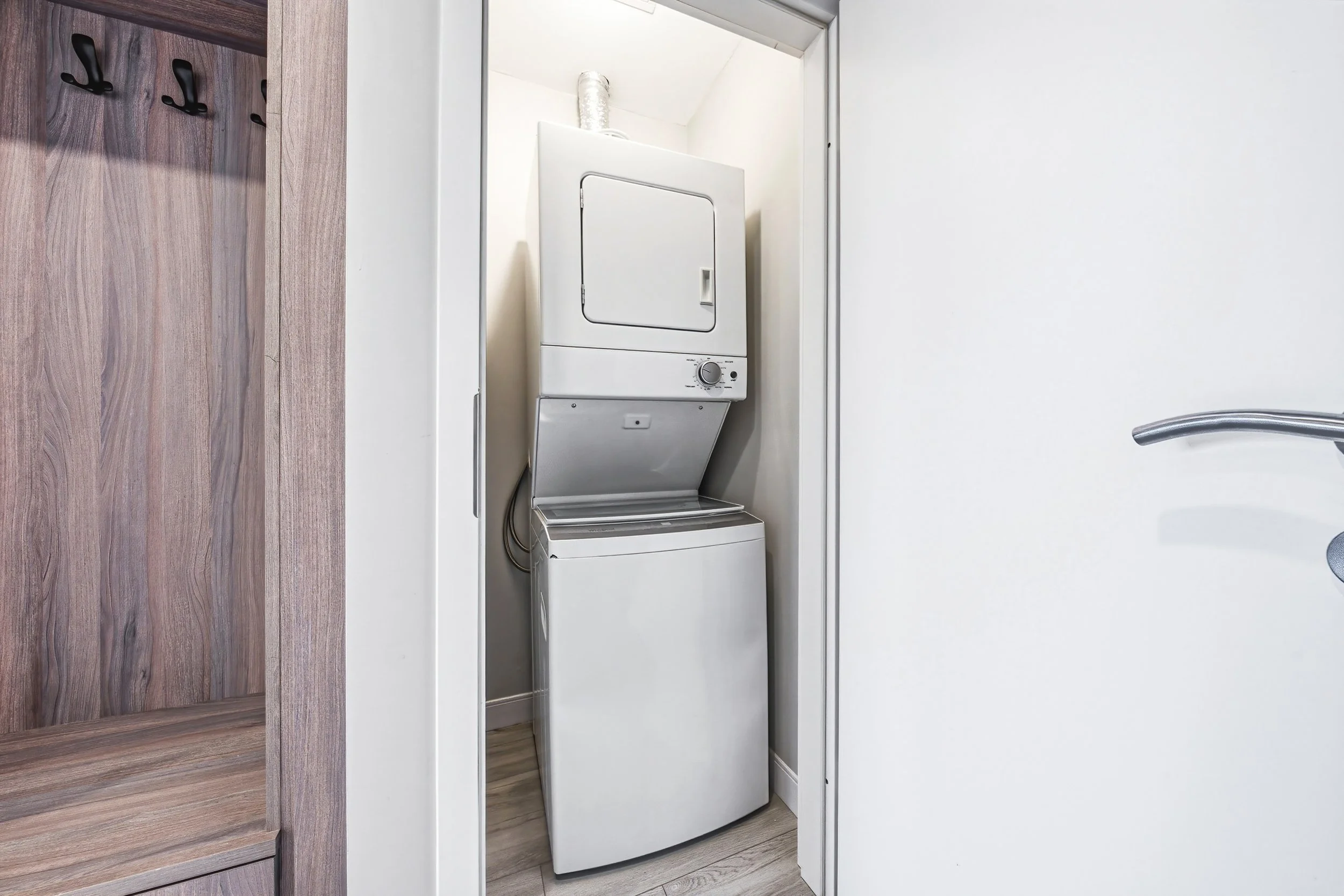 A laundry room with a stacked washing machine and dryer inside a small closet with a door frame. To the right, part of a silver door handle is visible, and to the left, wooden cabinetry with black hooks and woodgrain finish. The room has white walls 