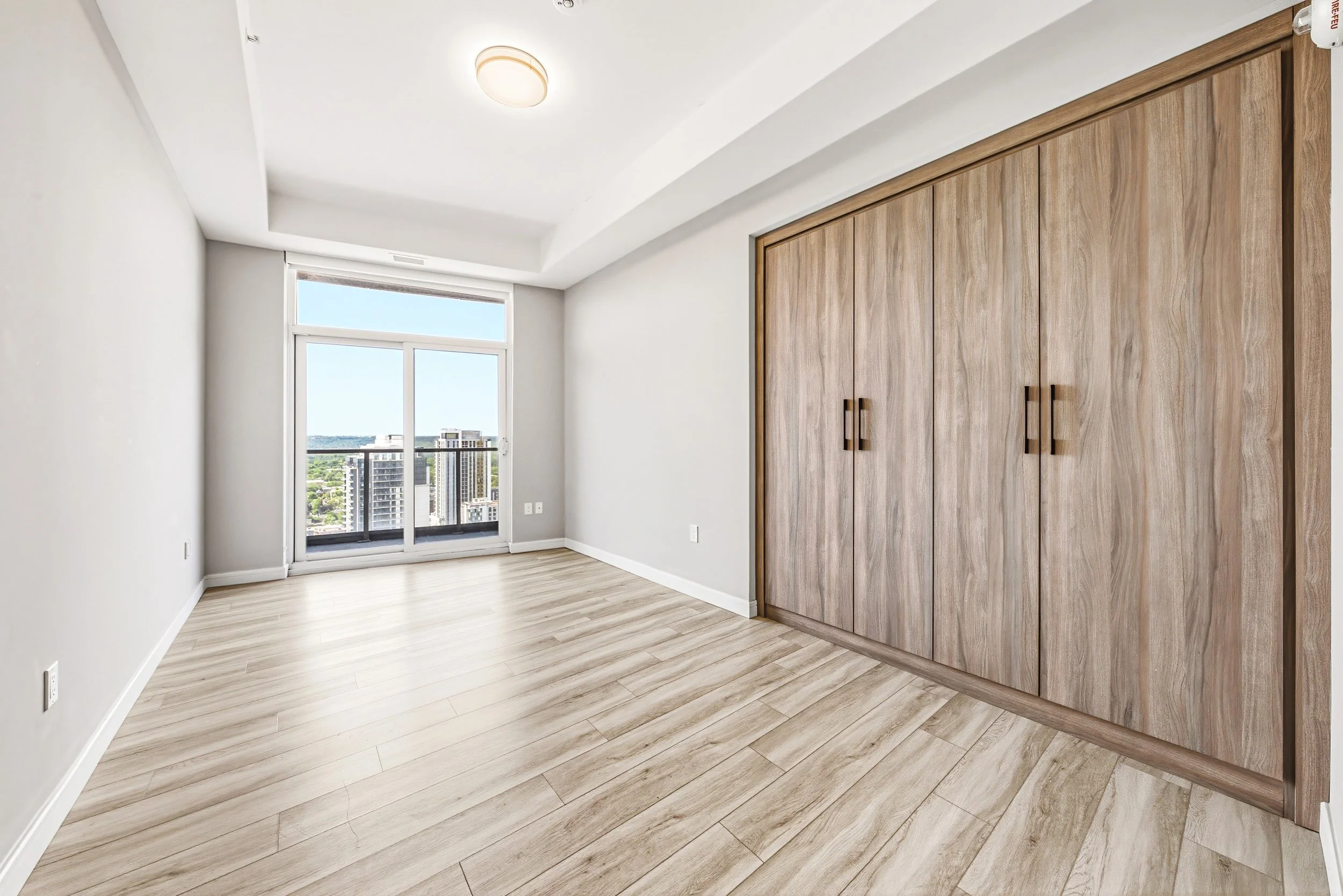 Empty room with wooden floors, white walls, a large window with a city view, and a built-in wooden closet.