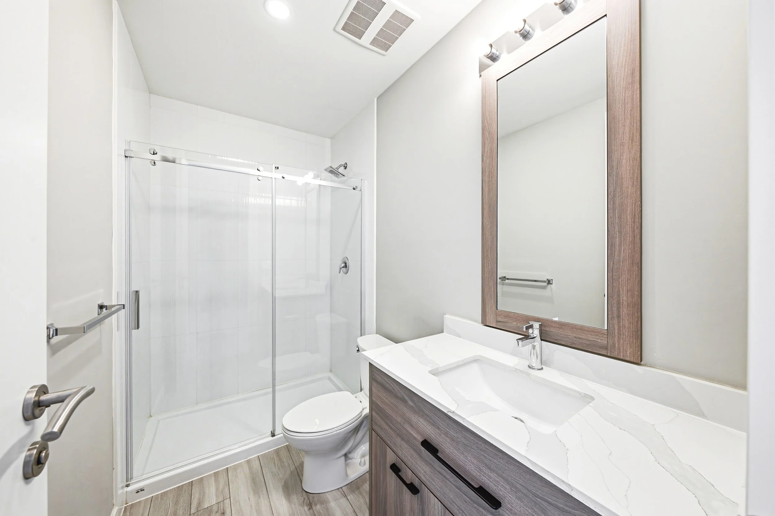 Modern bathroom with a walk-in shower with glass sliding doors, a white toilet, a white vanity with a marble top, a large mirror with a wooden frame, and wood-look tile flooring.