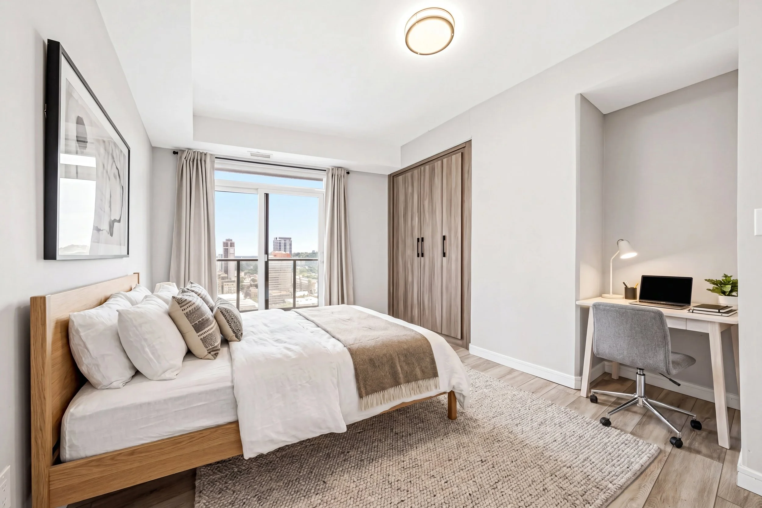Modern bedroom with a wooden bed, white bedding, pillows, a large window with city view, closet with wooden sliding doors, a small white desk with a gray office chair, lamp, and plant.