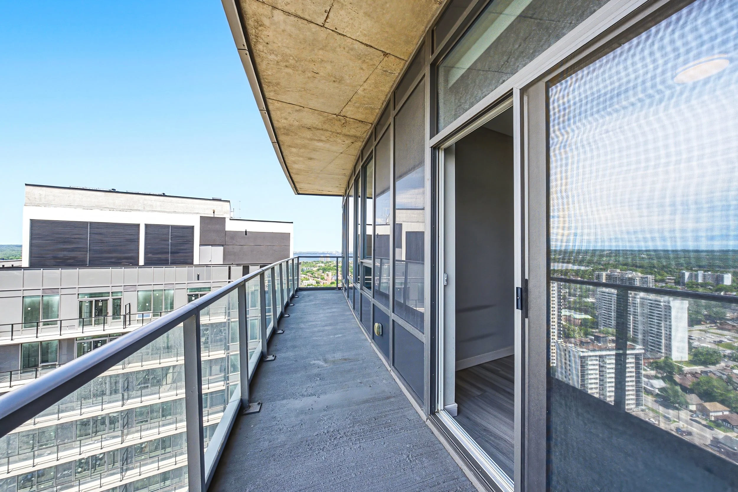 A view from a balcony of a high-rise building showing neighboring buildings, cityscape, and a blue sky. The balcony has glass railings, concrete floor, and sliding glass doors.
