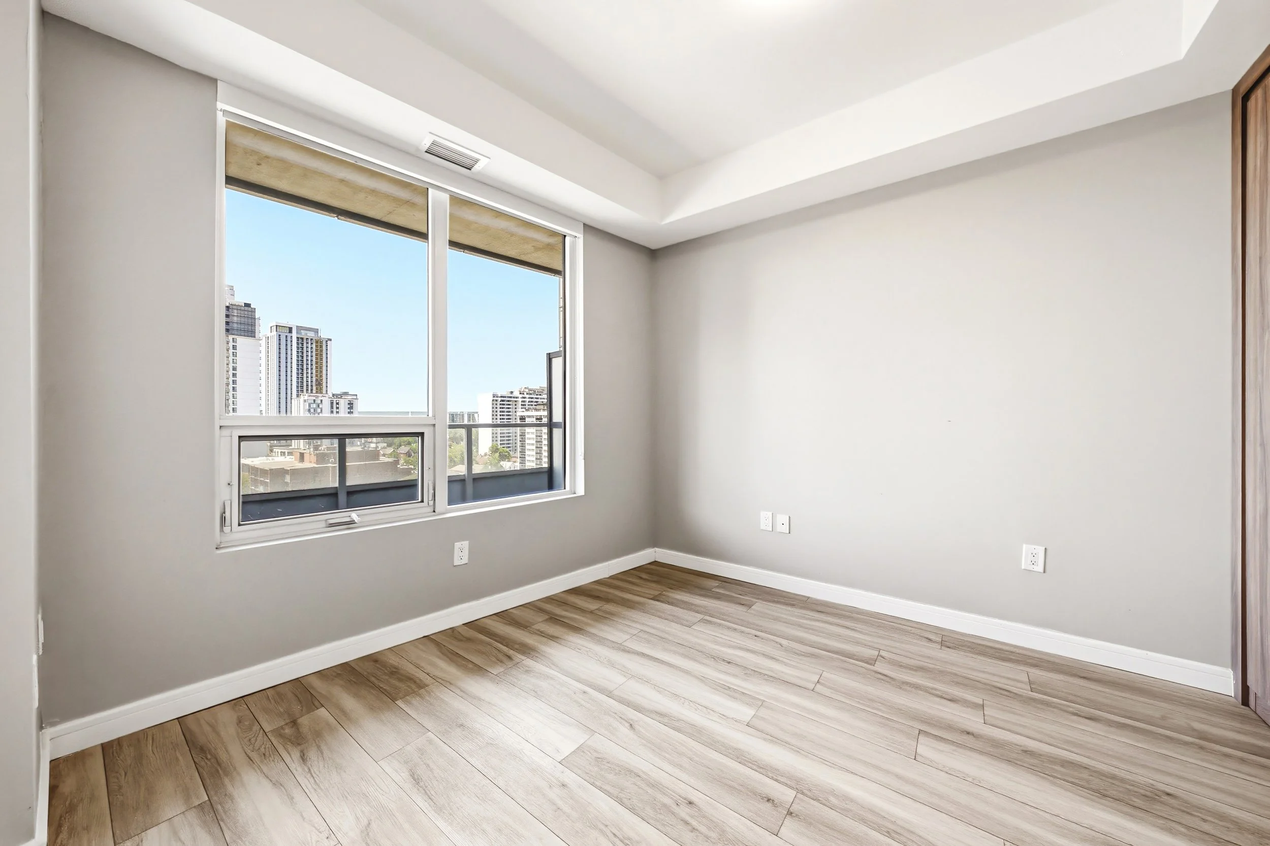 Empty room with hardwood floors and large windows showing city buildings outside.
