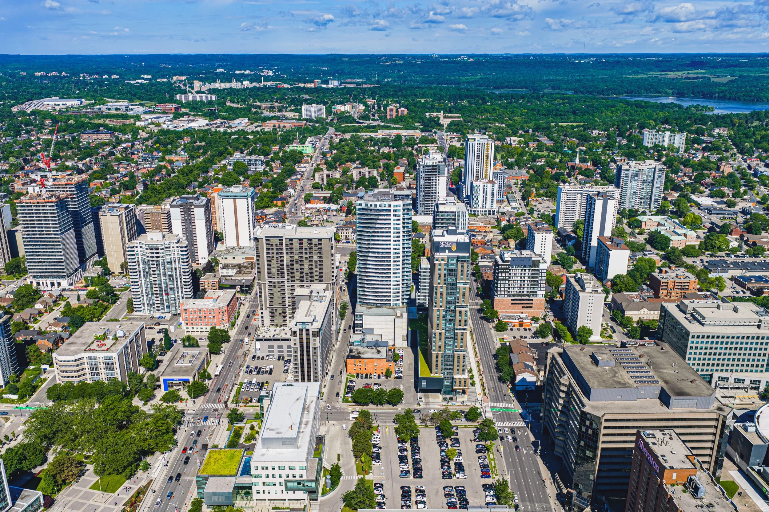 Aerial view of a city with tall modern buildings, streets, green trees, and a body of water in the background.