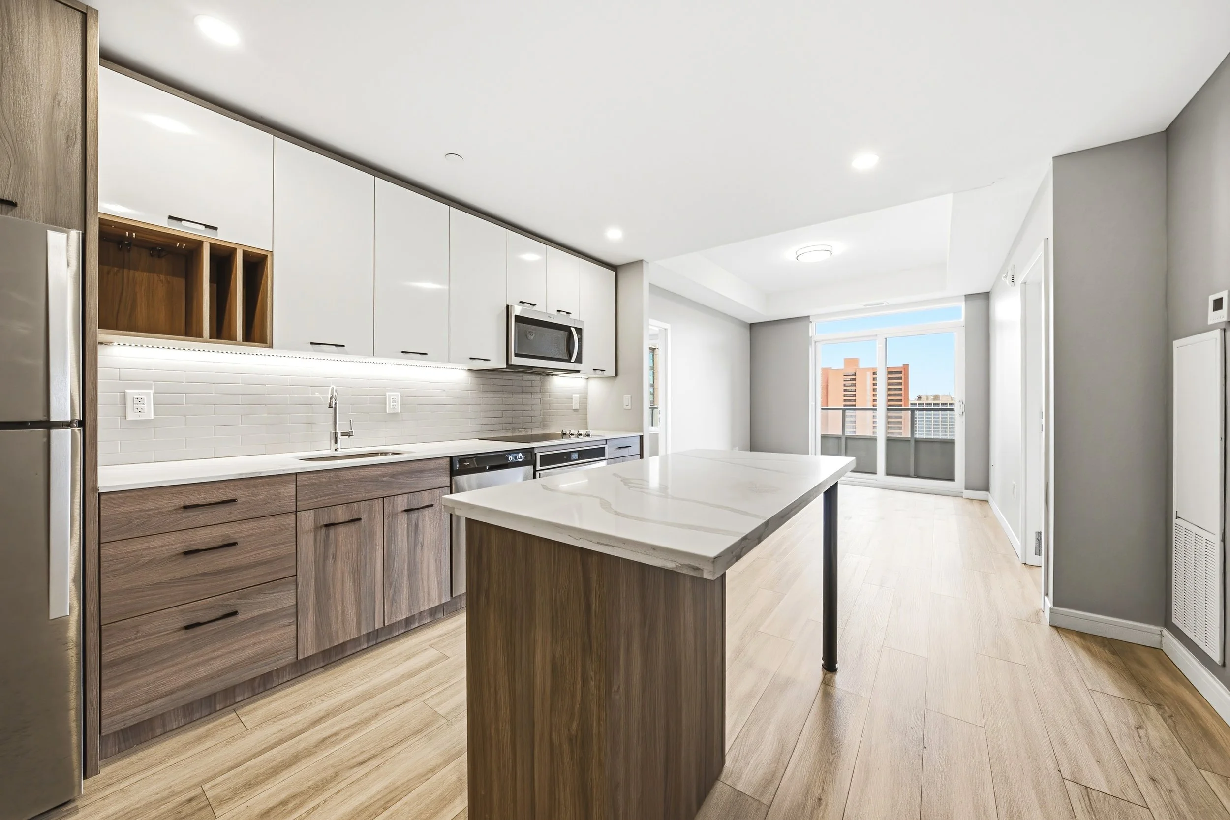 Modern kitchen with white upper cabinets, wooden lower cabinets, a marble countertop island, stainless steel appliances, and large windows providing natural light and city views.