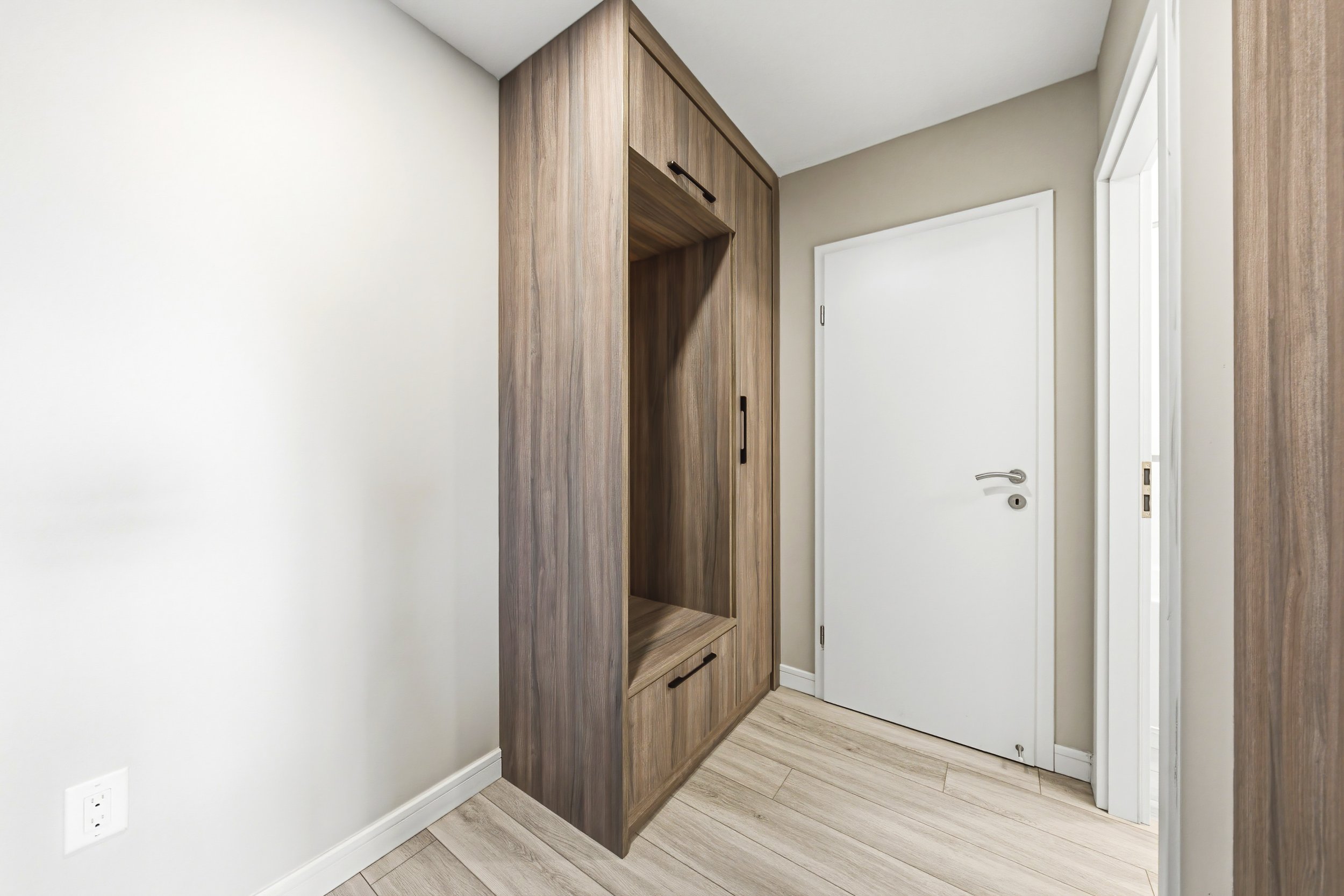 Interior of a room with light-colored walls, a wooden built-in cabinet with black handles, a white door with a silver handle, and light wood flooring.