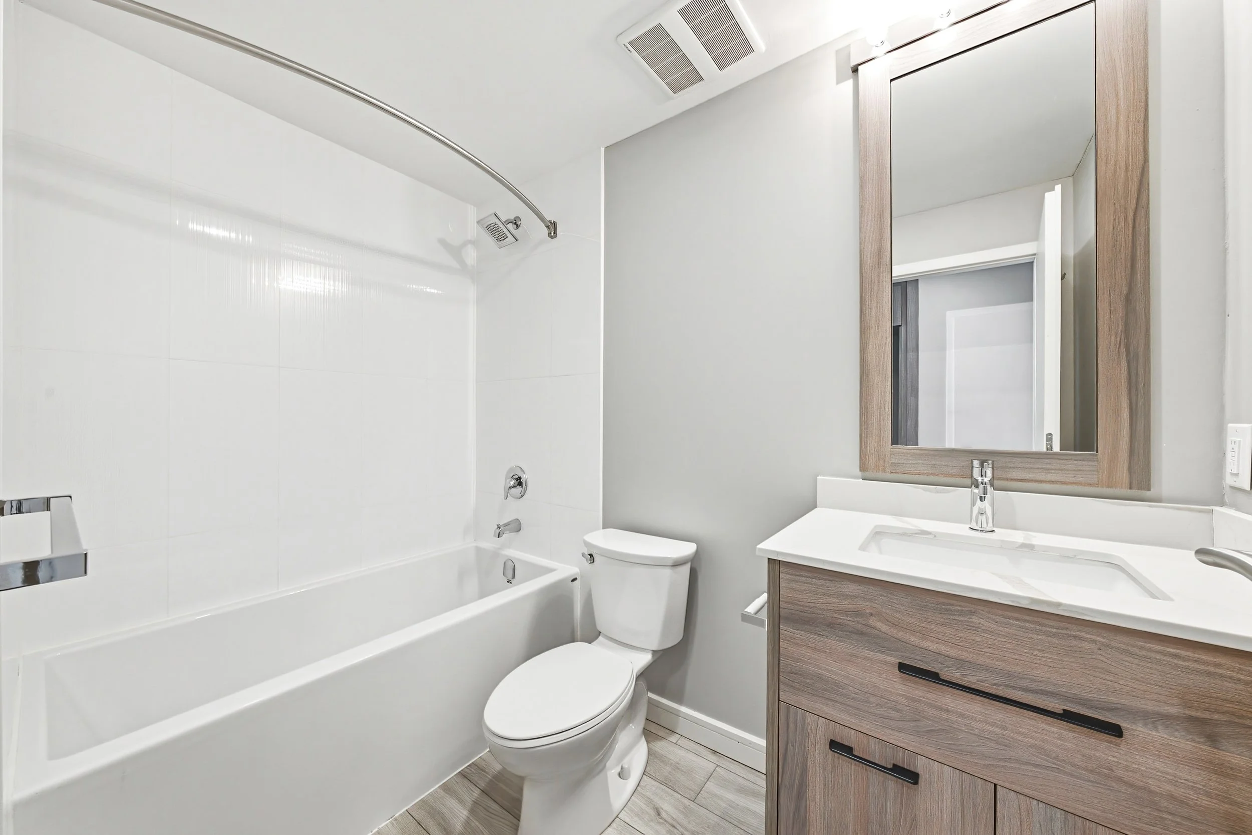 Modern bathroom with white bathtub, toilet, wooden vanity with white countertop and mirror, light-colored wood flooring, and deep gray walls. Fixtures include a showerhead, faucet, and cabinet handles.