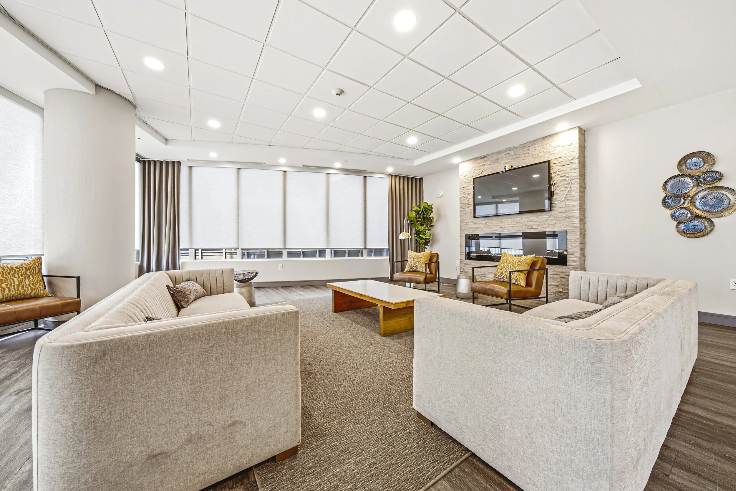 Modern living room with beige sectional sofa, yellow accent pillows, wooden coffee table, wall-mounted TV, stone fireplace, green plant, and large windows with blinds.