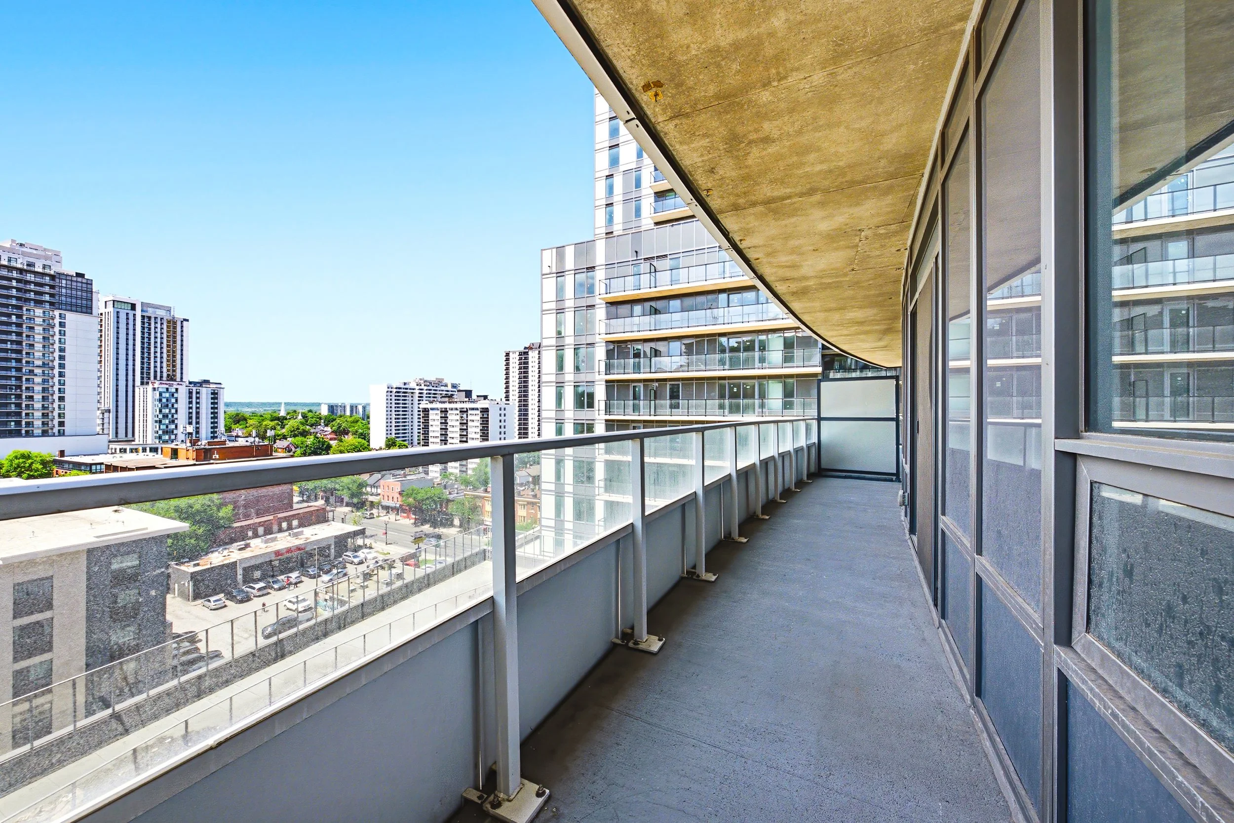 Empty balcony space with glass railing, view of high-rise buildings and parking lot, under clear blue sky.