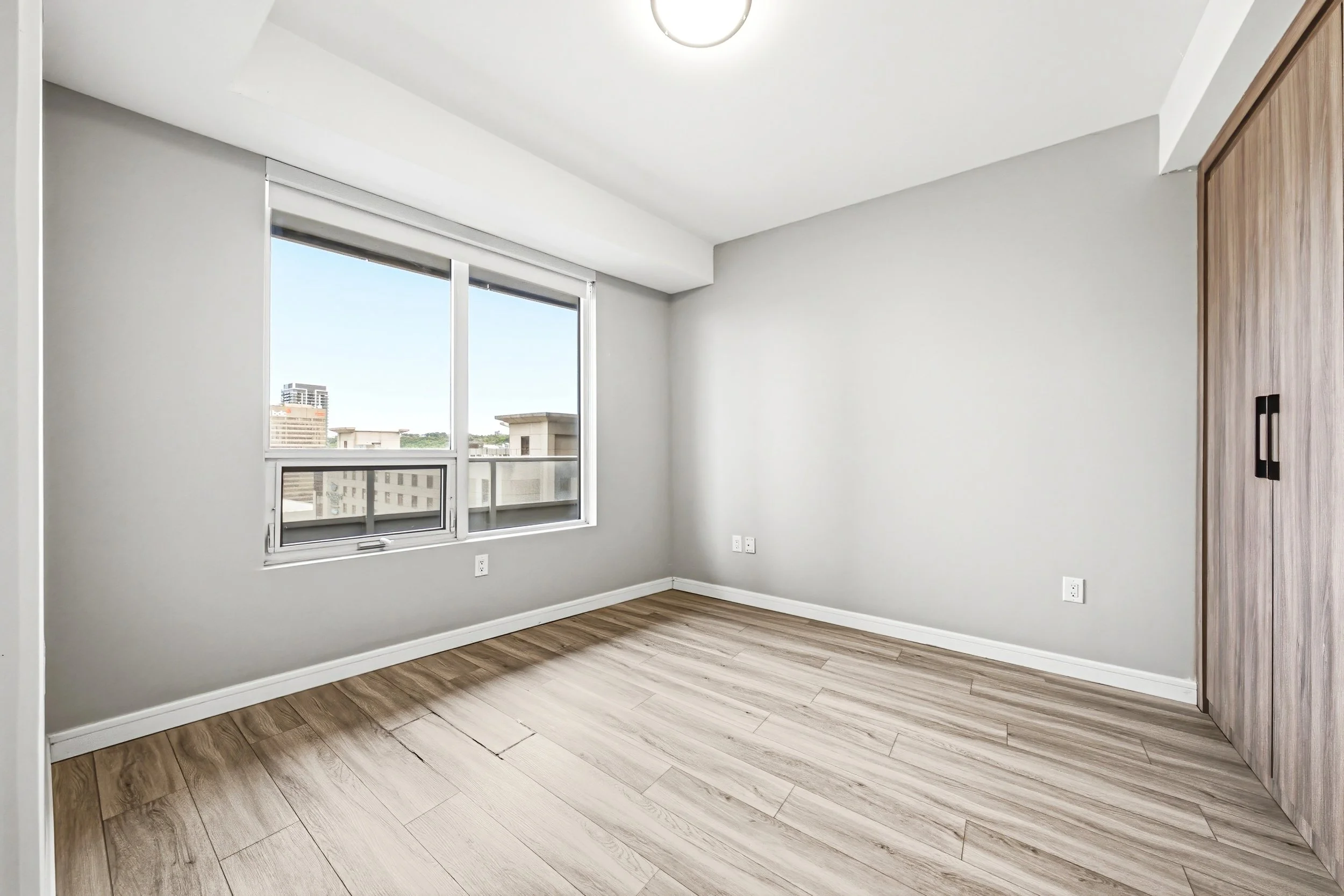 Empty room with large windows, white walls, wood flooring, and a wooden closet door.