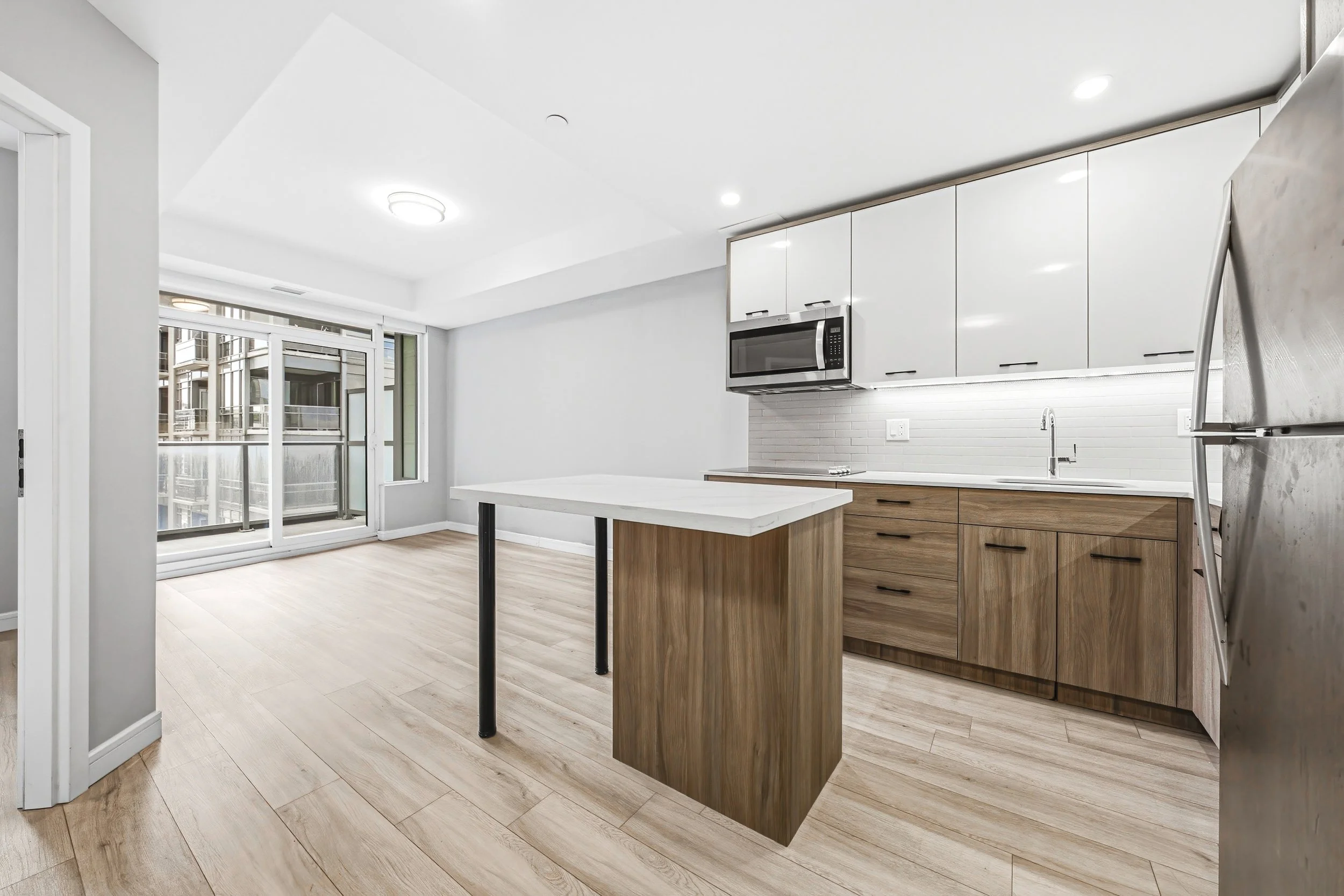 Modern kitchen with white upper cabinets, wooden lower cabinets, a microwave, stainless steel refrigerator, and a sliding glass door leading to a balcony.