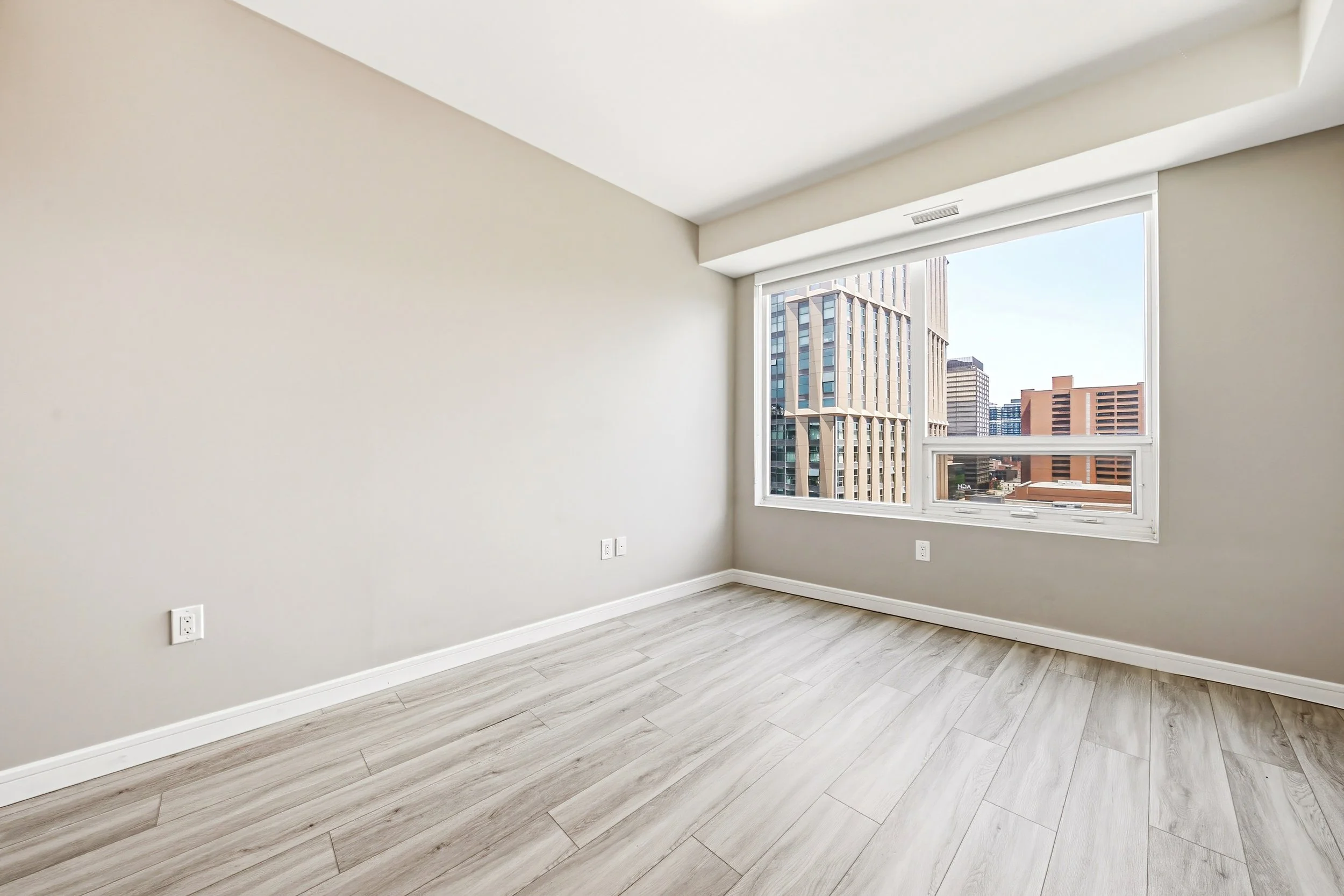 Empty room with large window showing a city skyline, beige walls, light wood flooring, and electrical outlets.