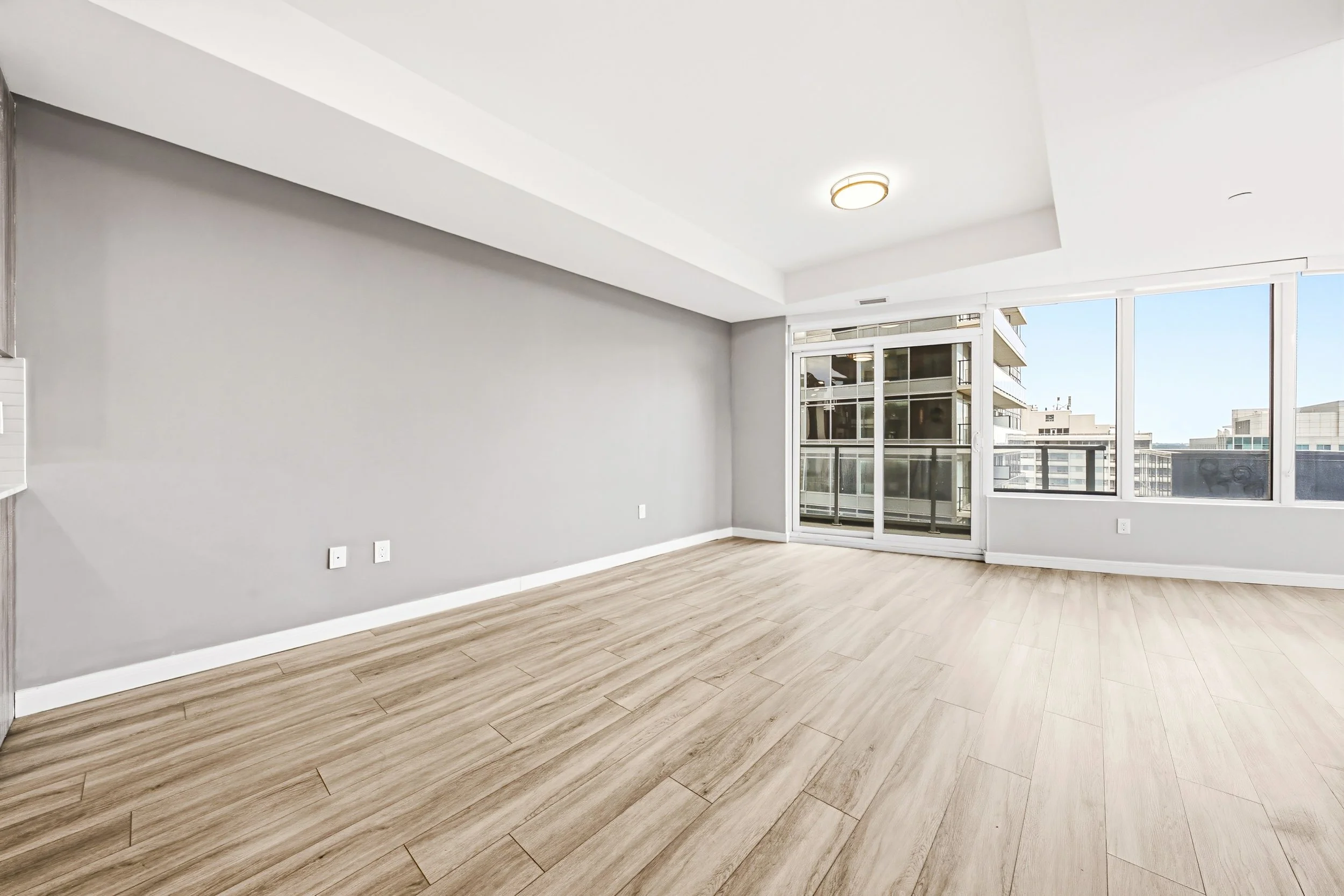 Empty living room with large windows, light wood flooring, and gray walls.