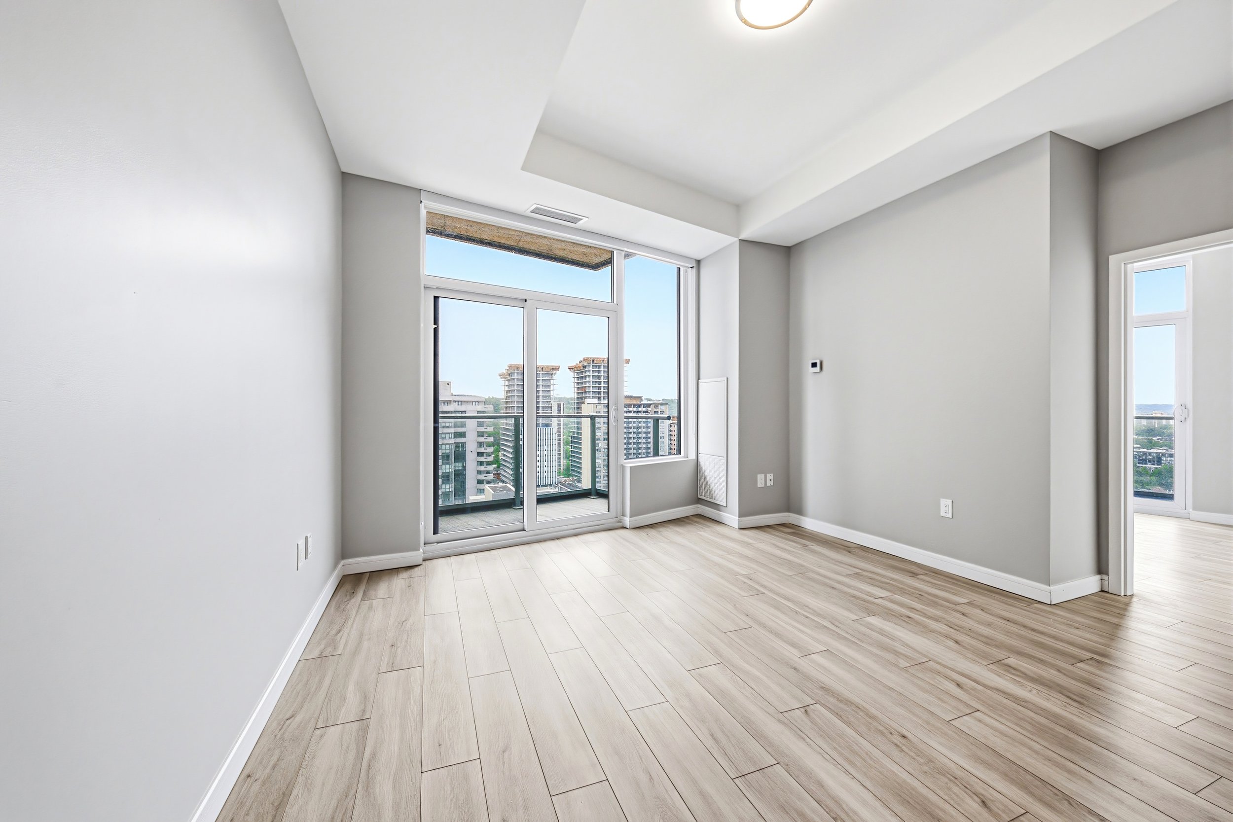 Empty modern apartment room with large windows and city view, white walls, light wood flooring.