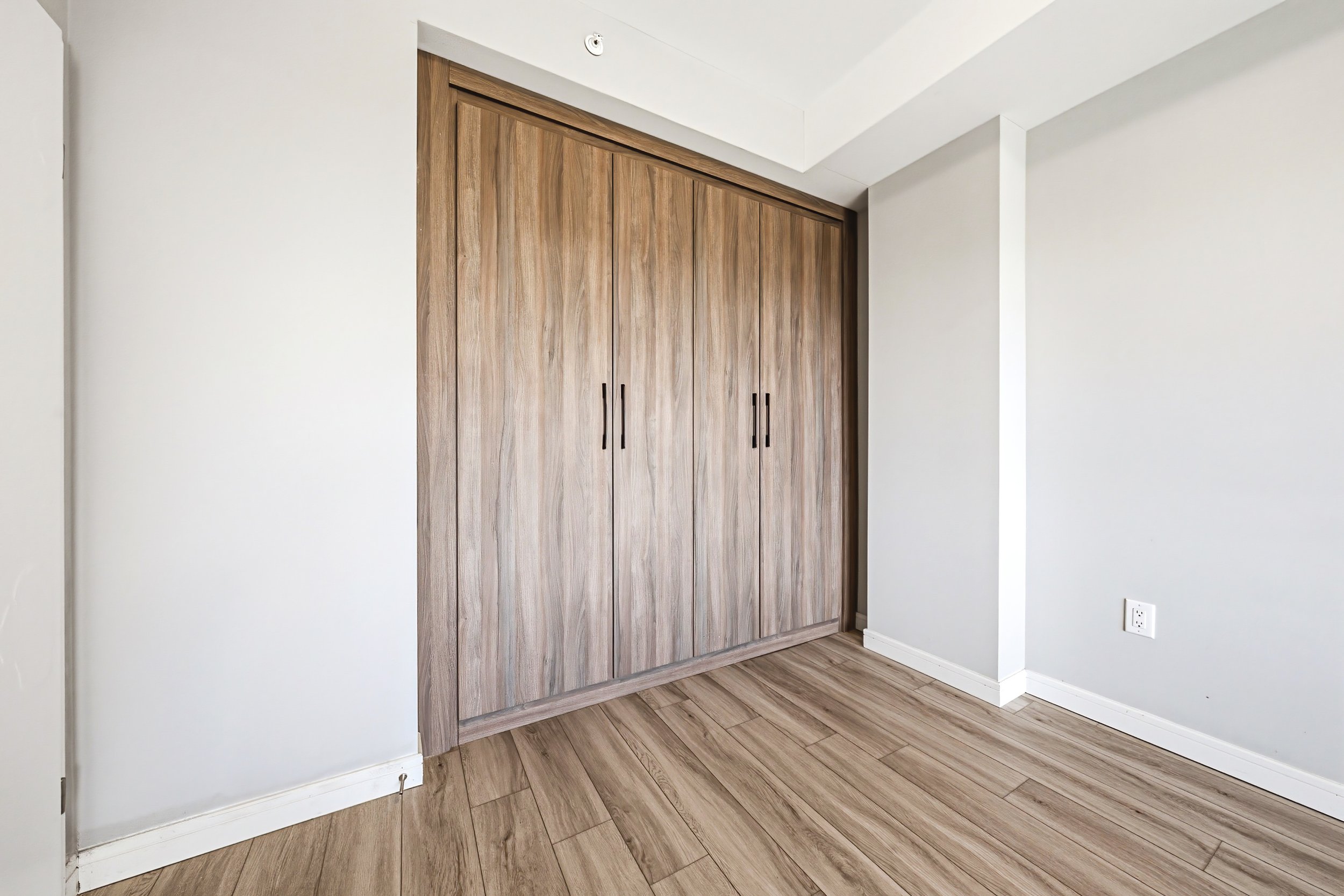 A closed wooden wardrobe with four black handles in a room with white walls and wooden floor.
