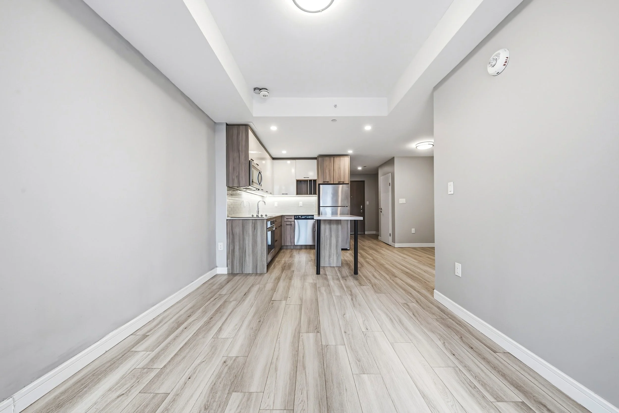 Empty living space with light wood flooring, white walls, and a view into a kitchen with wooden cabinets, stainless steel appliances, and a small kitchen island.
