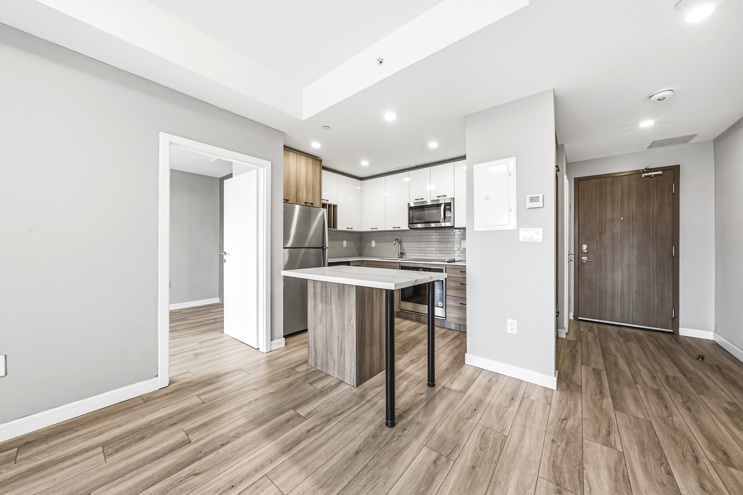 Modern kitchen with white cabinets, stainless steel appliances, a kitchen island with a wooden base, and wood-look flooring. Visible area includes an entrance door and part of the living space.