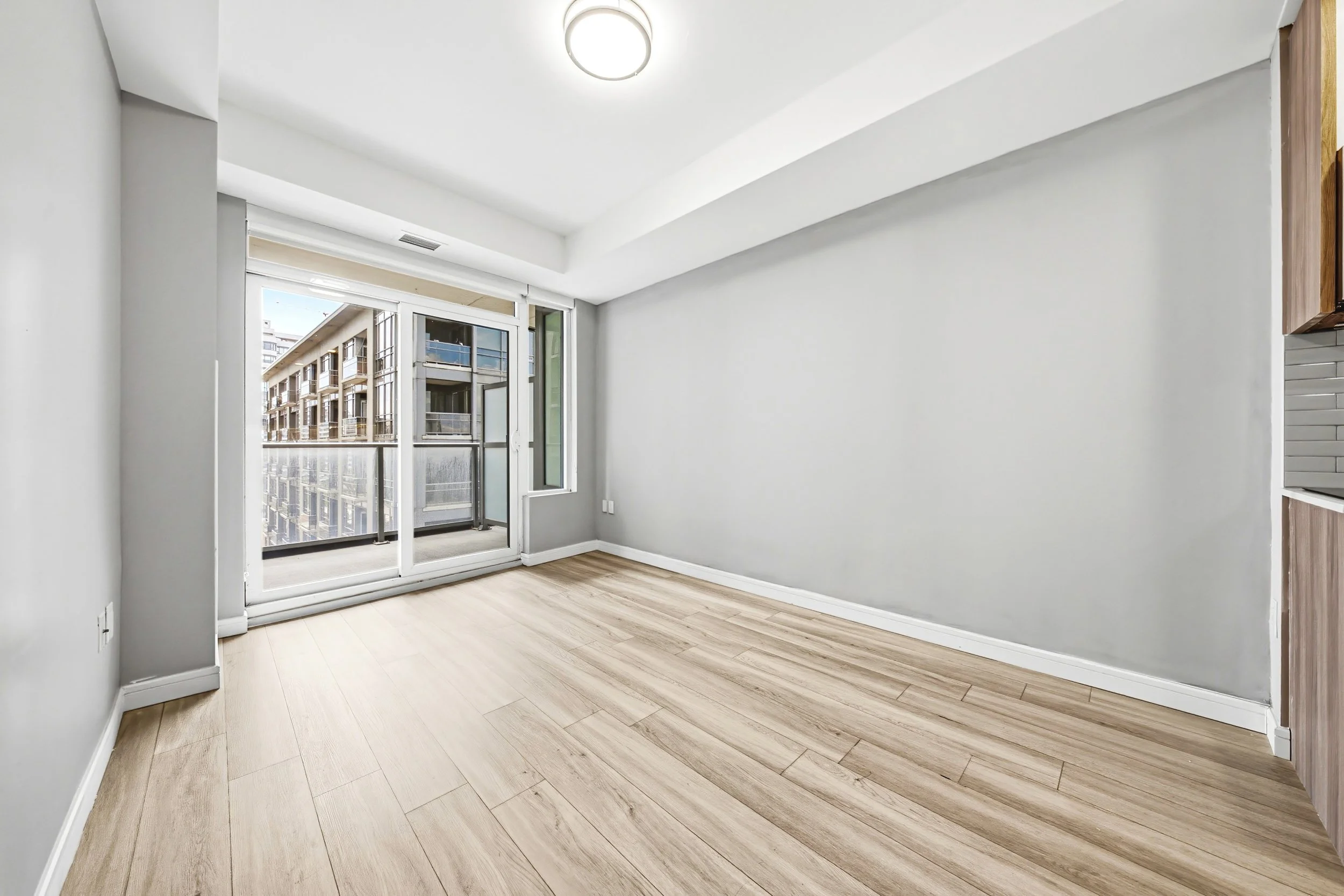 Empty room with light-colored wood flooring, white walls, and a large glass door leading to a balcony overlooking neighboring buildings.