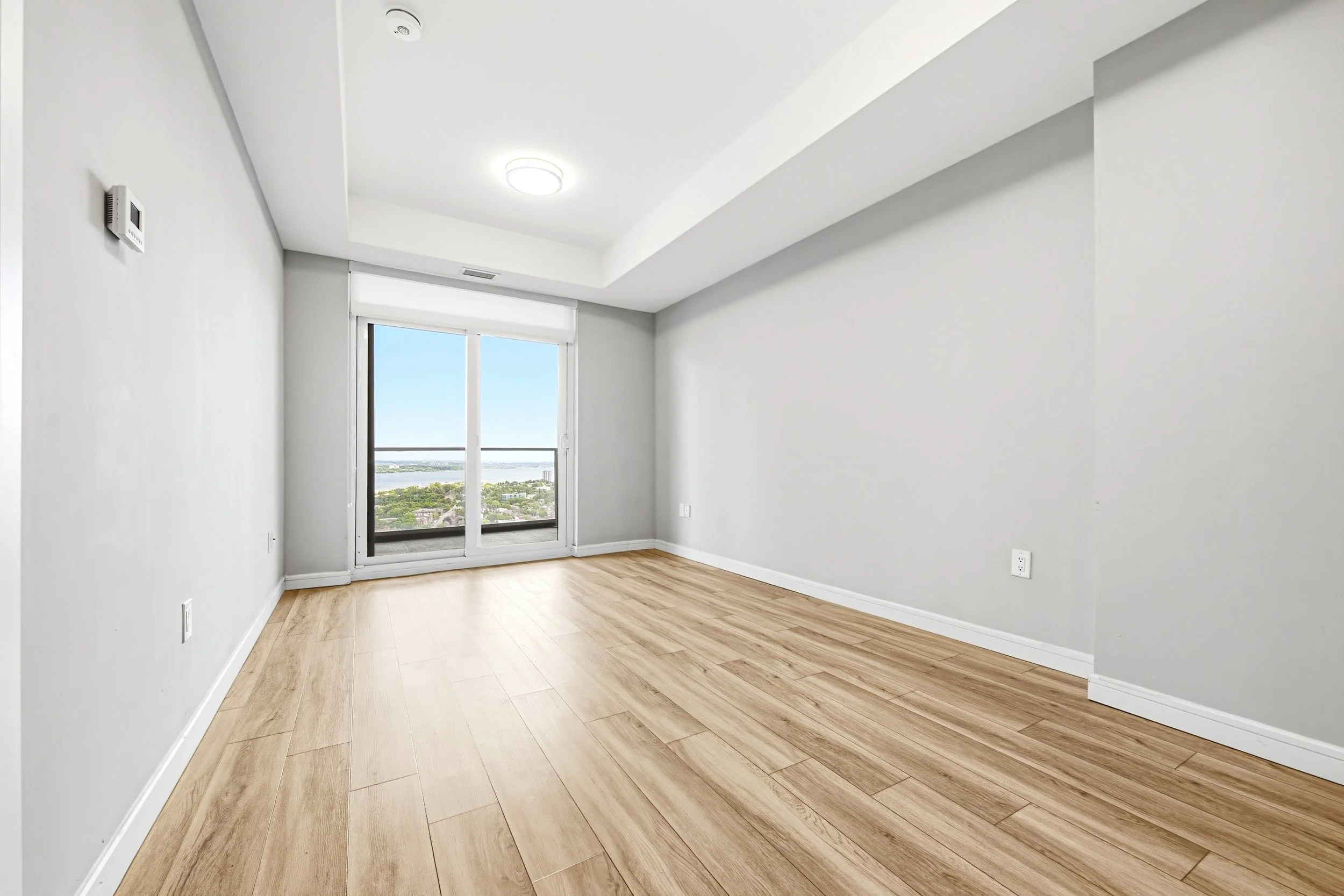 Empty room with white walls, light wood flooring, and a large sliding glass door leading to a balcony with a view of trees and water.