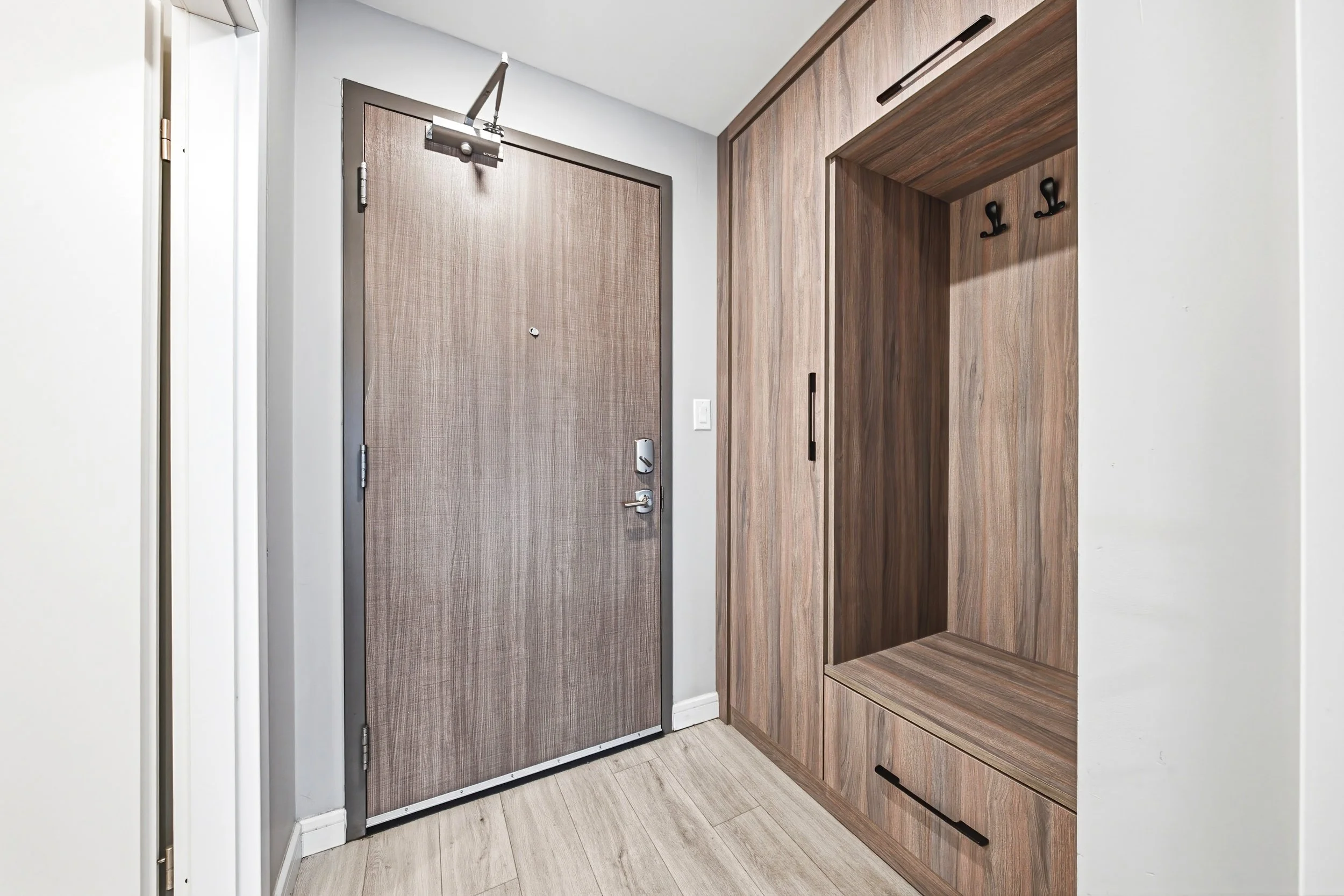 Interior view of a hallway showing a gray door with a door closer and lock, and a built-in wooden closet with black hooks on the side.