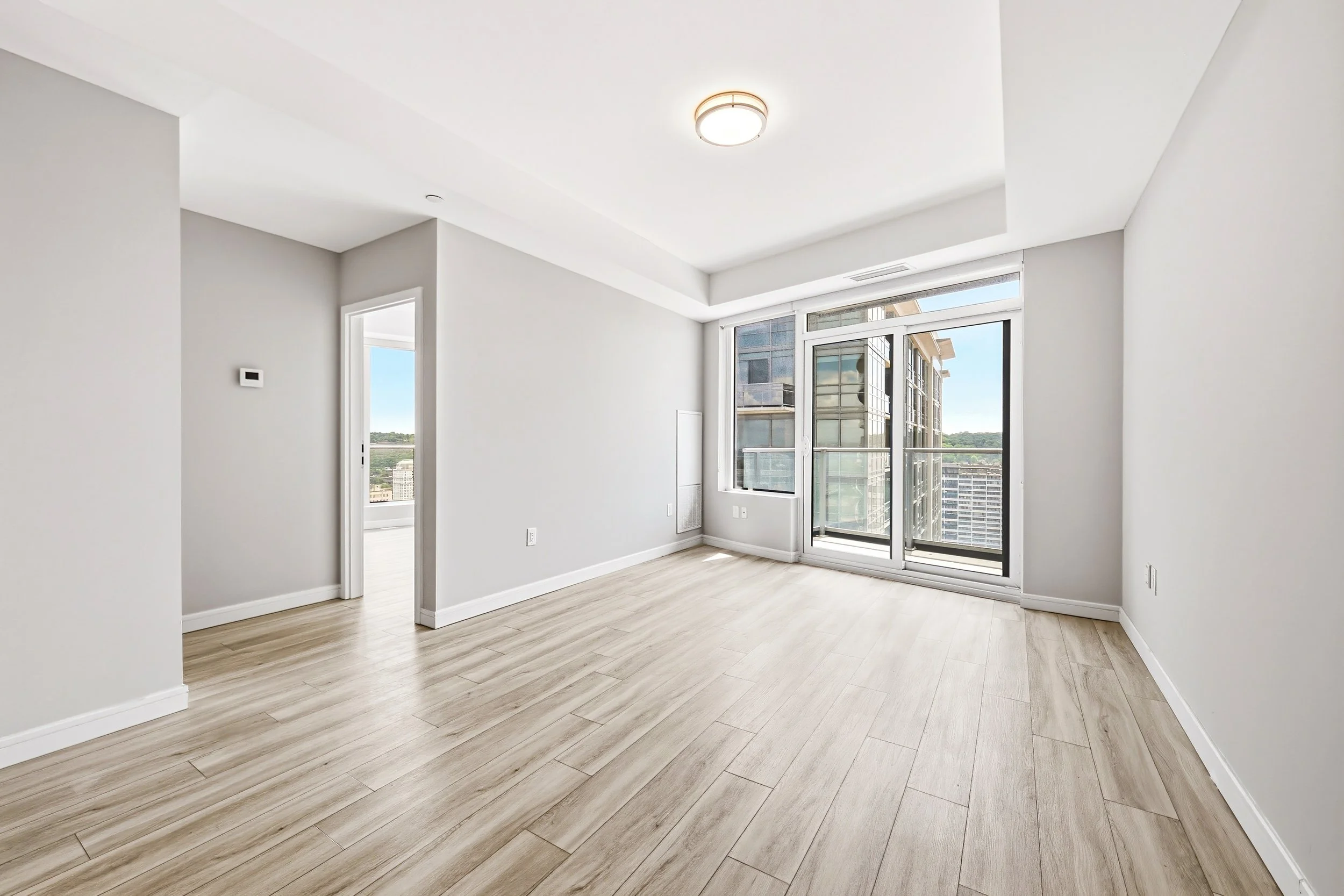 Empty modern apartment room with large window showing city buildings, light wood flooring, white walls, and ceiling light fixture.