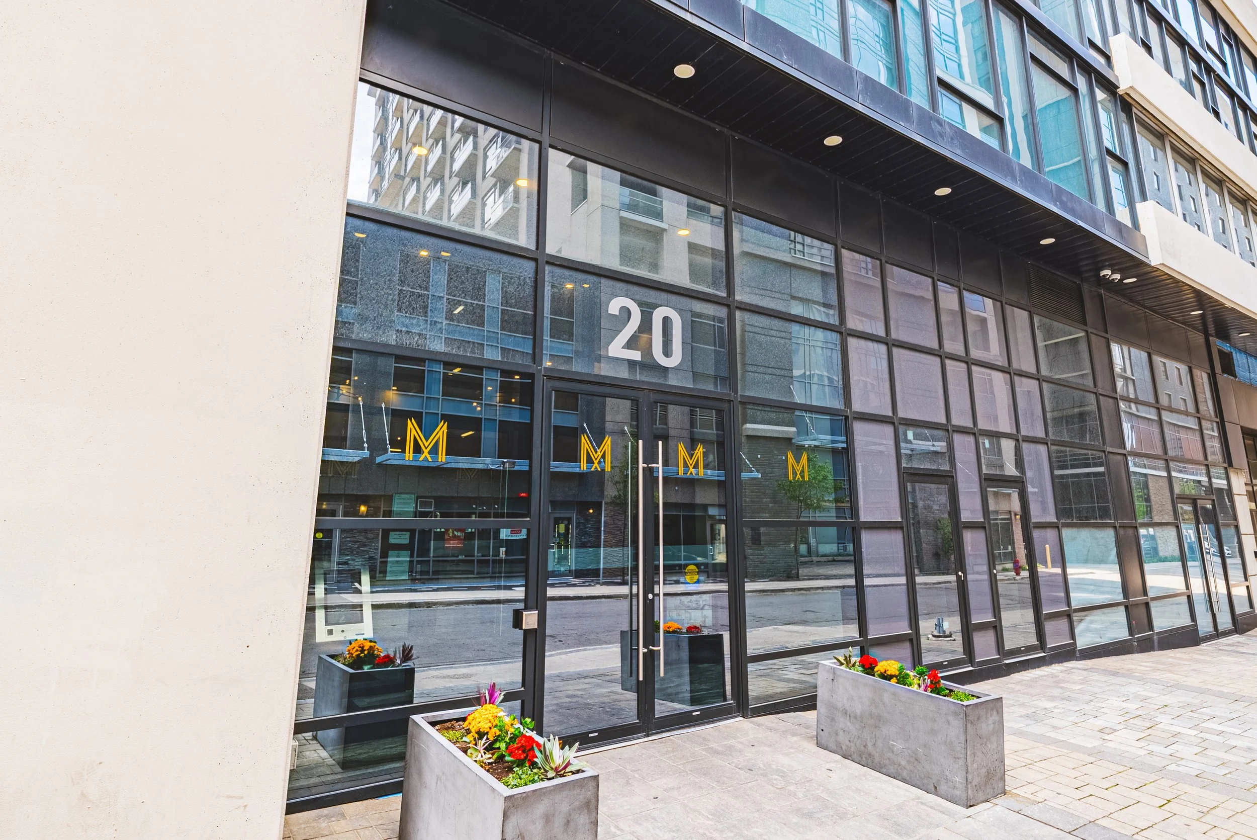 Modern glass building with the number '20' displayed above entrance, surrounded by flower planters, with city buildings reflected in windows.