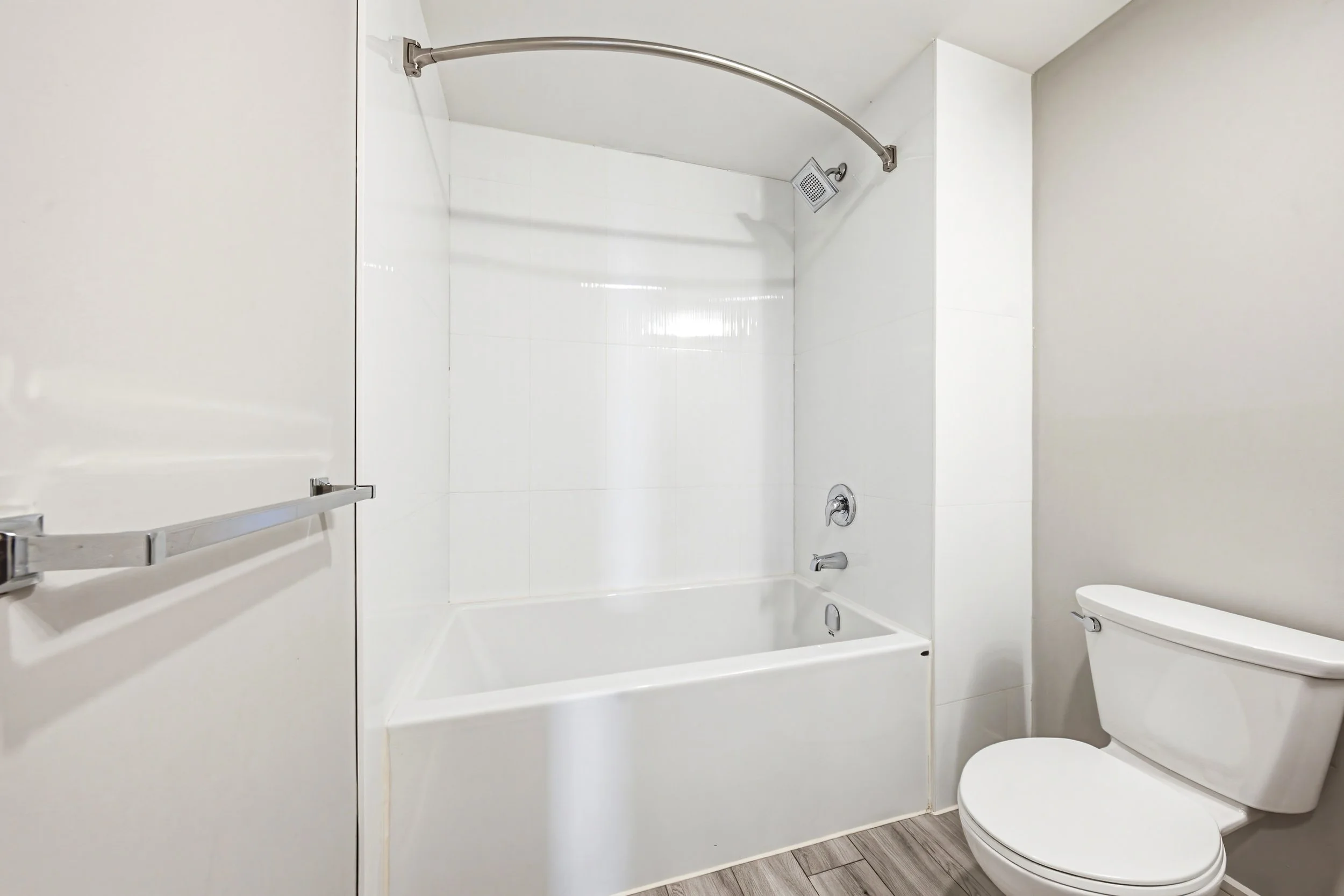 Modern bathroom featuring a bathtub with a shower, a glass shower door, a handheld showerhead, a toilet, and a pull-up bar mounted on the wall, with white walls and wood-patterned floor.